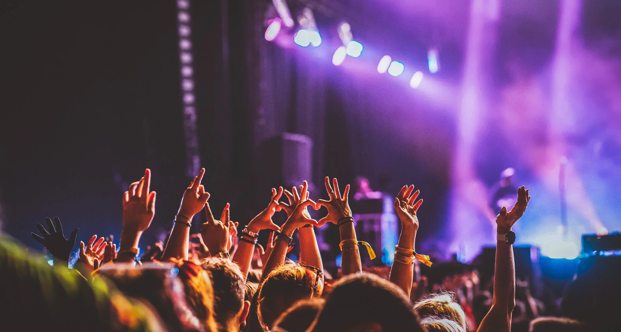 Crowd at a concert with many hands raised, some making heart shapes and peace signs, with a stage and purple and blue lighting in the background.