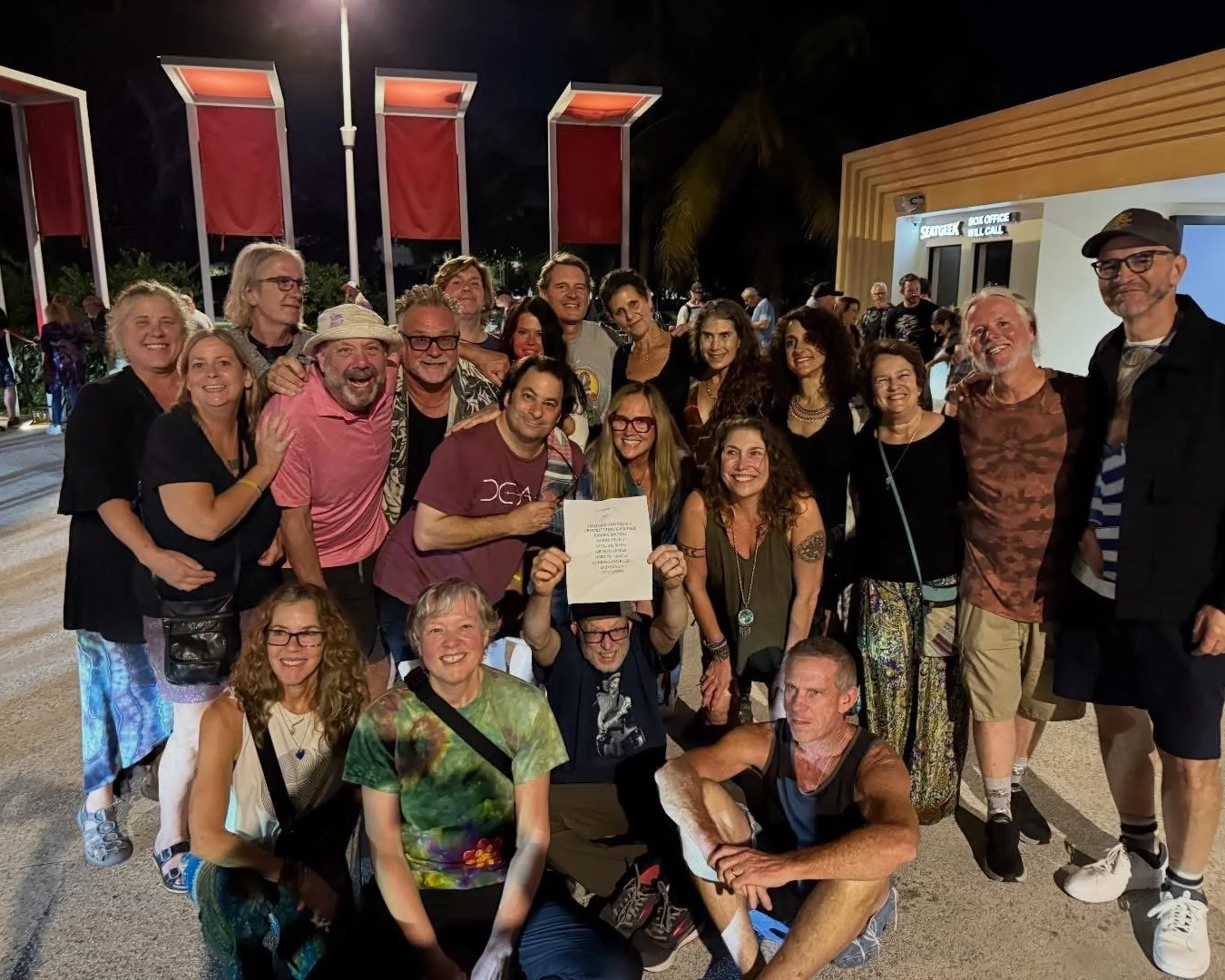 A large group of people smiling and posing together outdoors at night, some holding a piece of paper or certificate, in front of a building with a sign reading 'SECURITY, BOOKSTORE, MAIL CELL'.