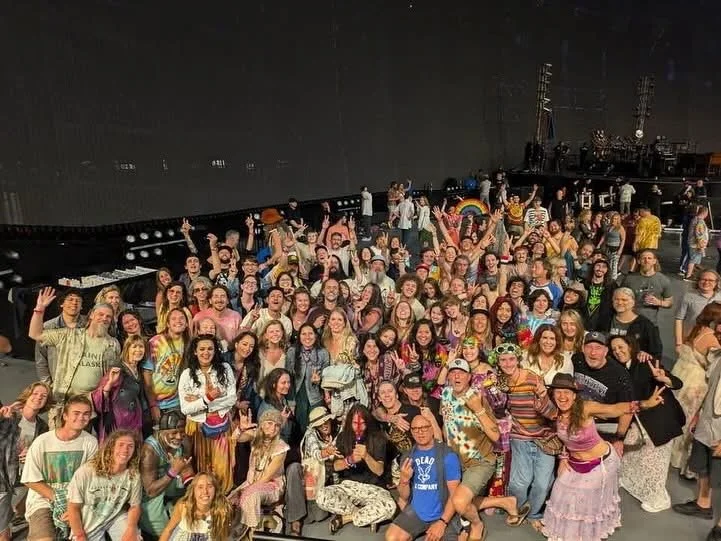 Large group of people gathered on stage or in an indoor venue, smiling and waving at the camera, with stage equipment and lighting in the background.