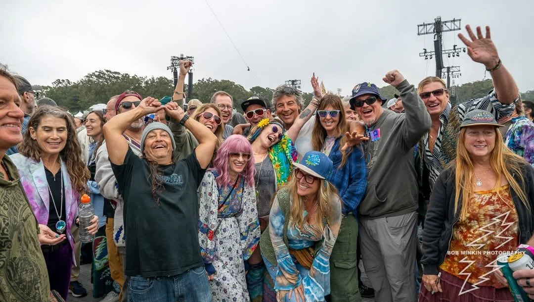 Group of people celebrating outdoors at a festival or event, smiling and cheering, wearing colorful and casual clothing, some with sunglasses, and raising their hands in excitement.