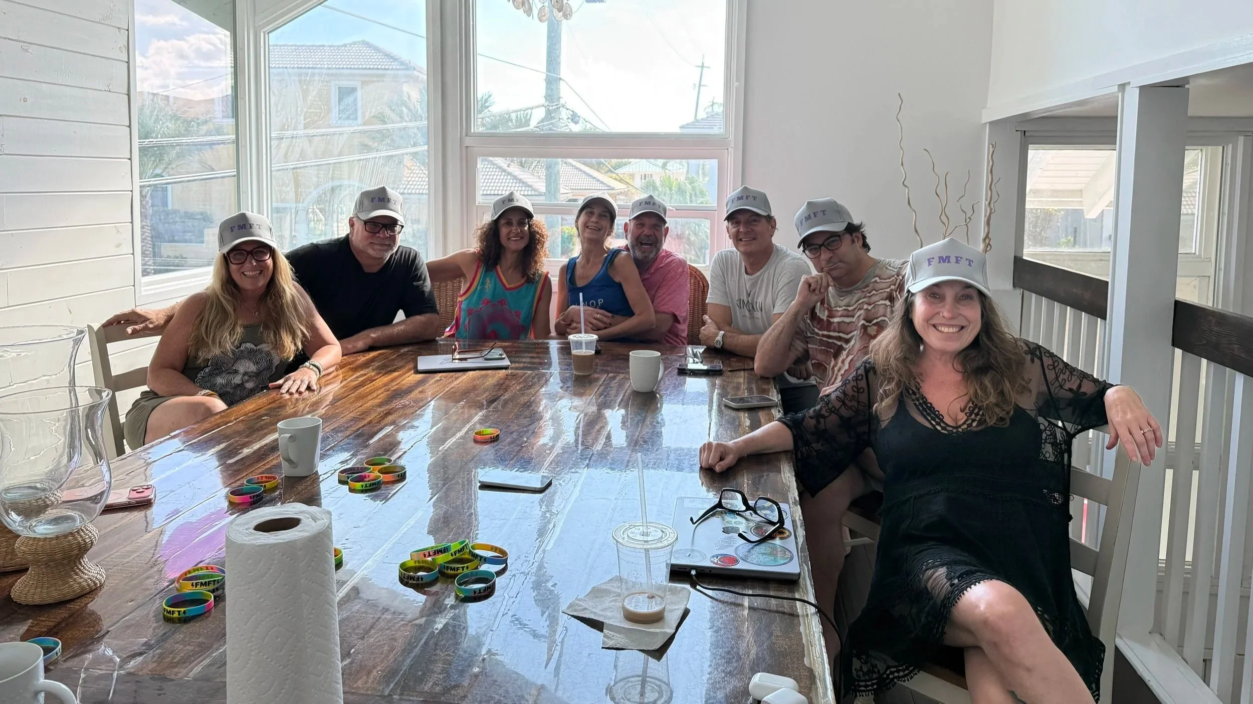 Group of eight people sitting around a large wooden table, wearing white caps with purple text, in a bright room with large windows, with some rings and cups on the table.