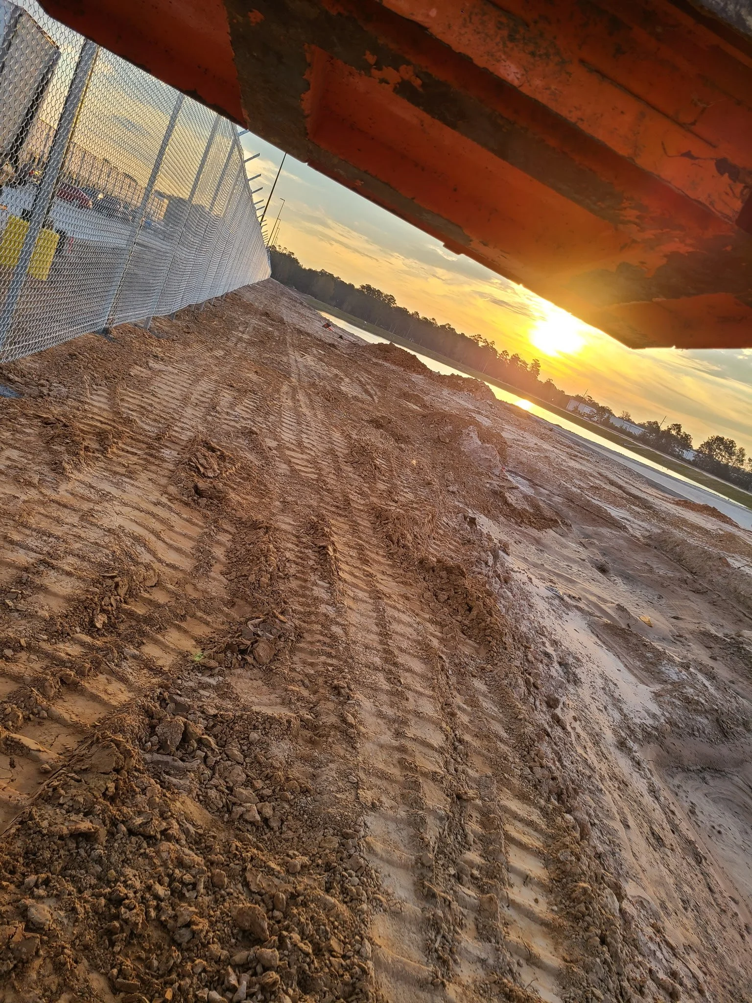 Construction site with dirt and tire tracks, metal fence on the left, and a large metal structure overhead. The sunset in the background over a body of water and trees.