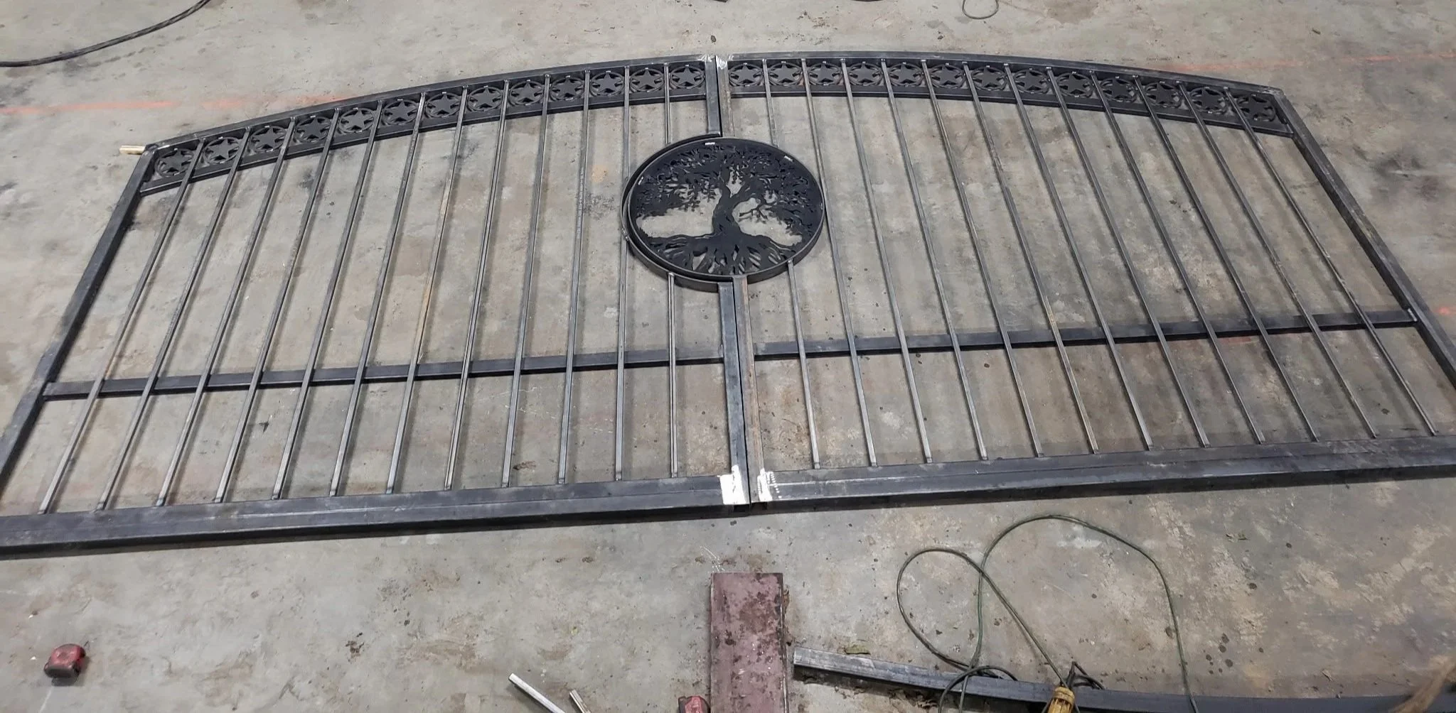 A large decorative metal gate with a tree emblem in the center lying on a concrete floor, surrounded by tools and materials.