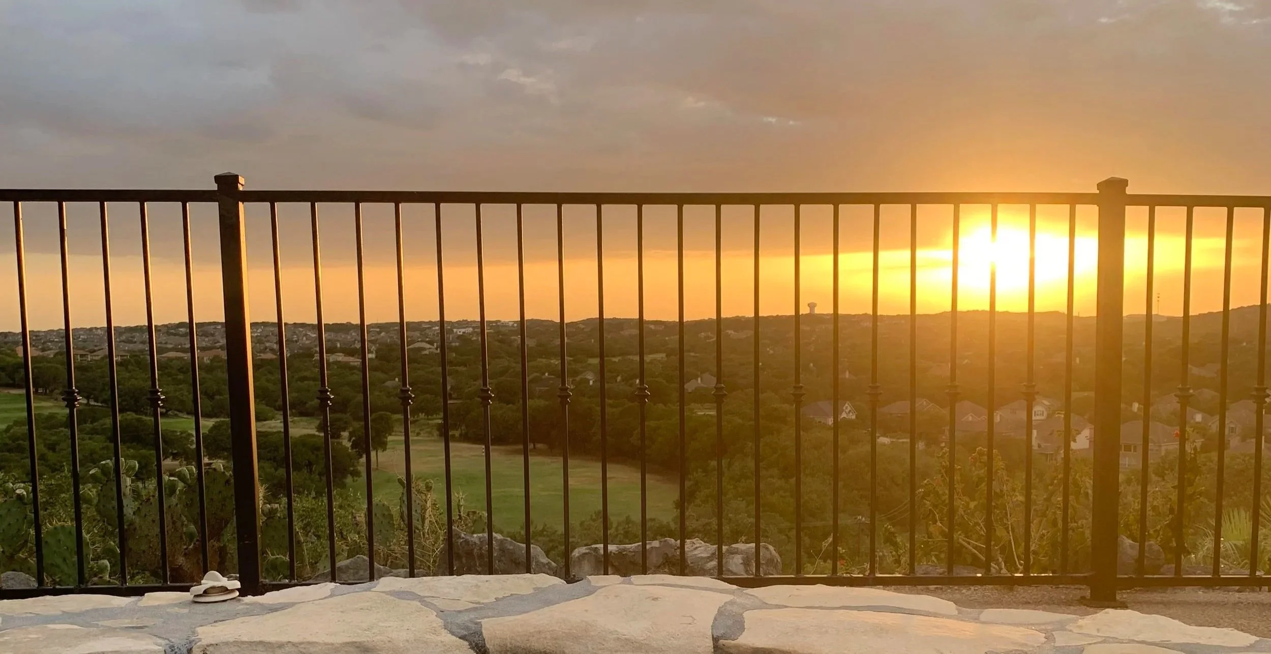 Sunset view over a landscape of trees, houses, and open fields, seen through a black metal railing with a stone patio in the foreground.