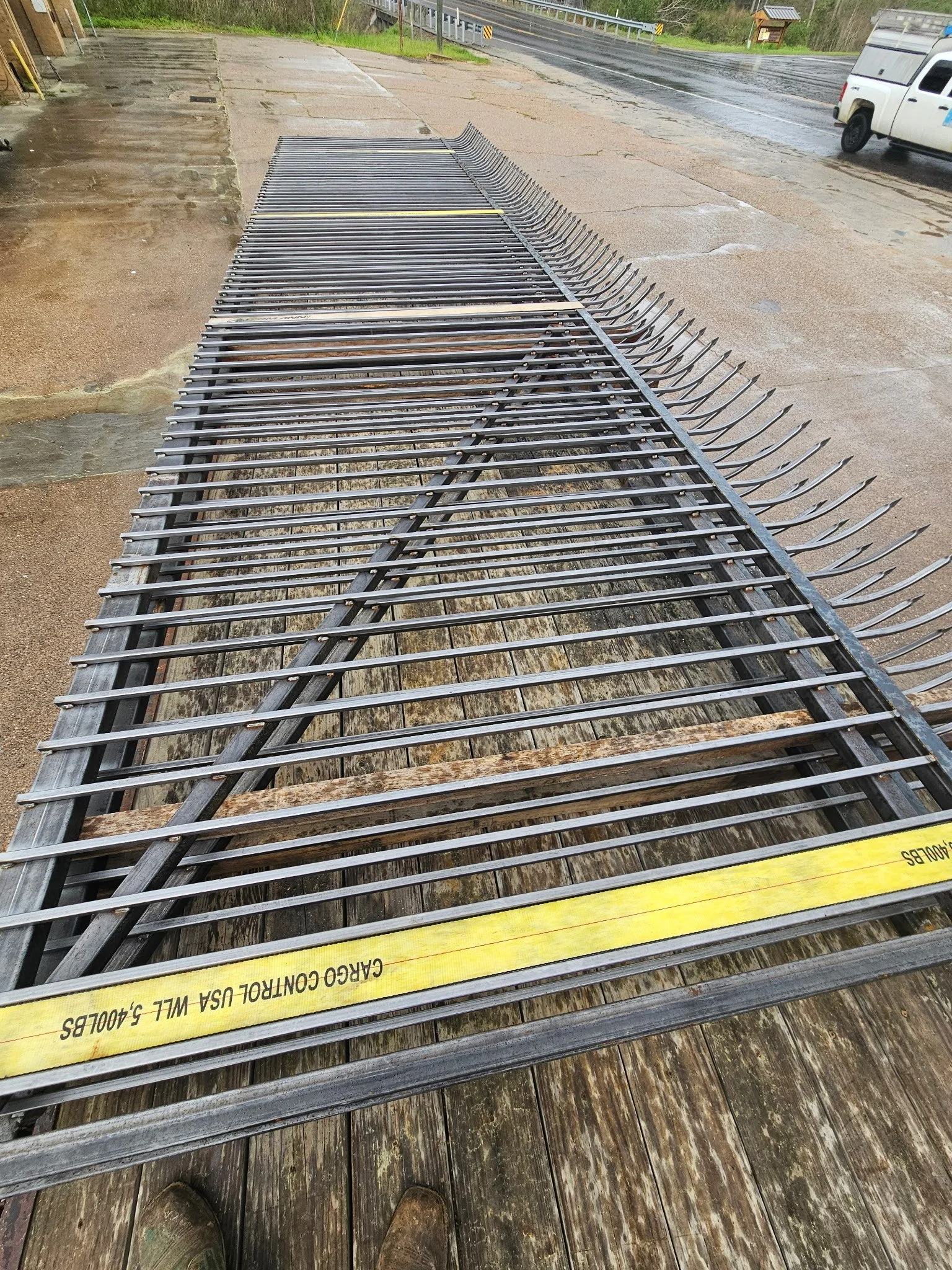 Metal guardrail sections stacked on a wooden platform outdoors on a rainy day near a roadway with a white utility truck in the background.