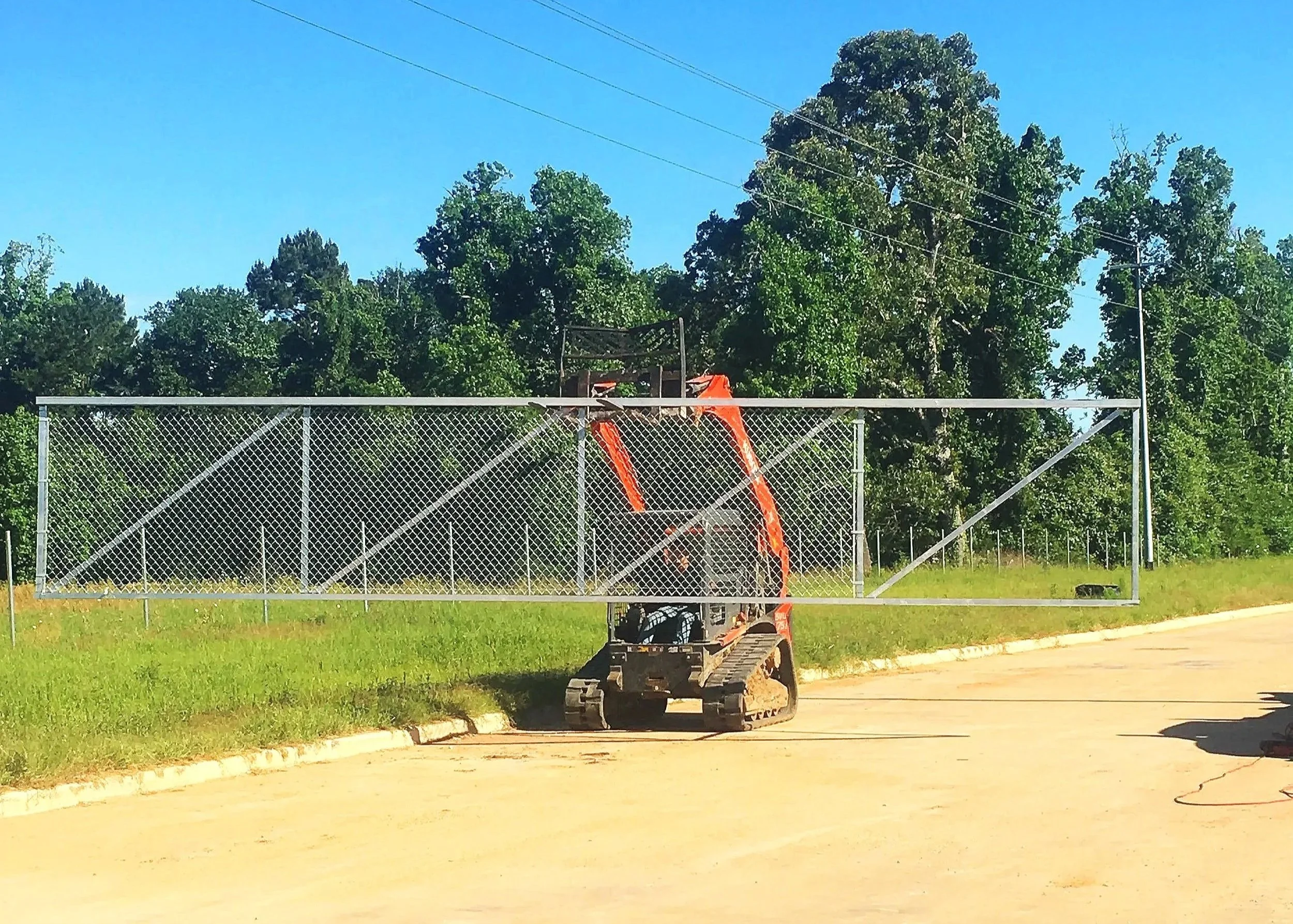 Construction vehicle working on installation of a chain-link fence along a paved road, with trees and a blue sky in the background.