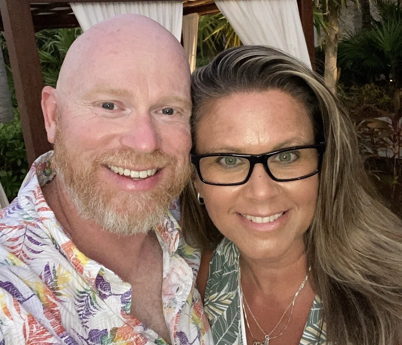 A man and woman taking a close-up selfie outdoors. The man has a bald head, light complexion, reddish beard, and is wearing a colorful, patterned shirt. The woman has long, wavy hair, glasses, and is wearing a similar patterned top with layered necklaces. There are plants and a wooden structure with curtains in the background.