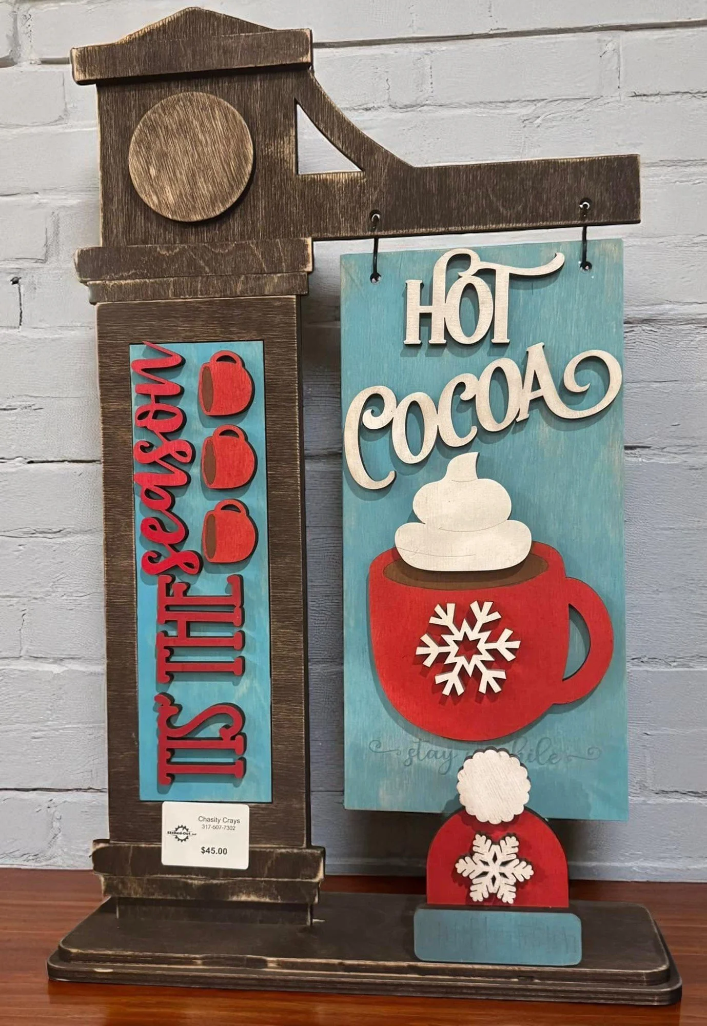 A decorative sign with a winter theme, featuring a red mug with a snowflake design, a white whipped topping, and a red and white snowflake decoration. The sign reads "Hot Cocoa" in large white letters, with additional text "Stay Cozy" in script. It is made of painted wood and displayed on a dark wooden base.