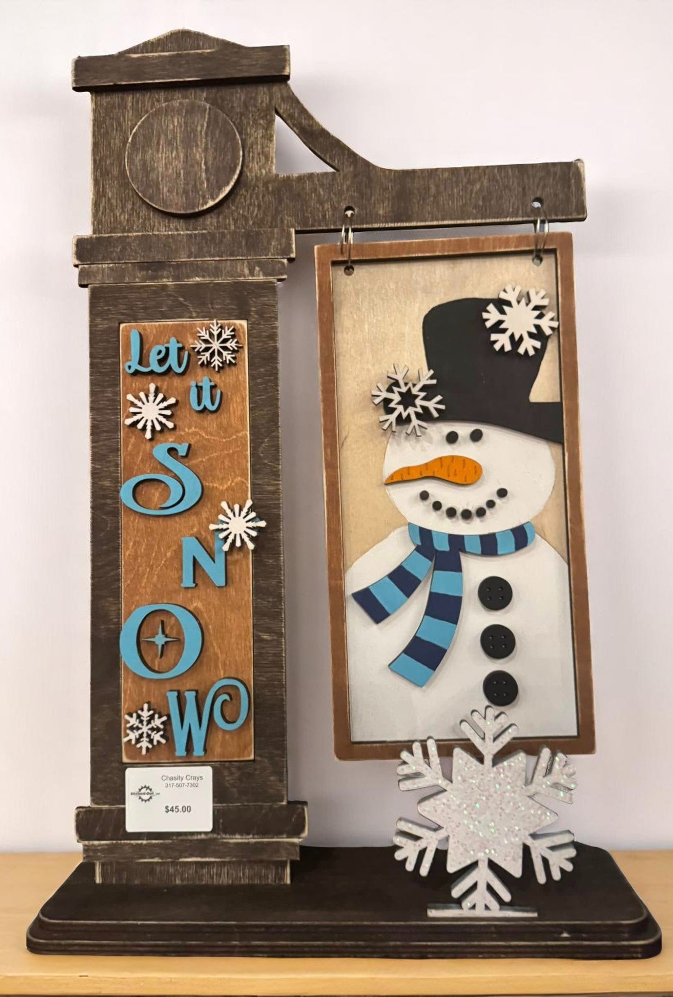 Decorative holiday sign featuring a snowman with a black hat, orange carrot nose, and blue scarf, with snowflake accents and the phrase 'Let it Snow' in colorful letters.