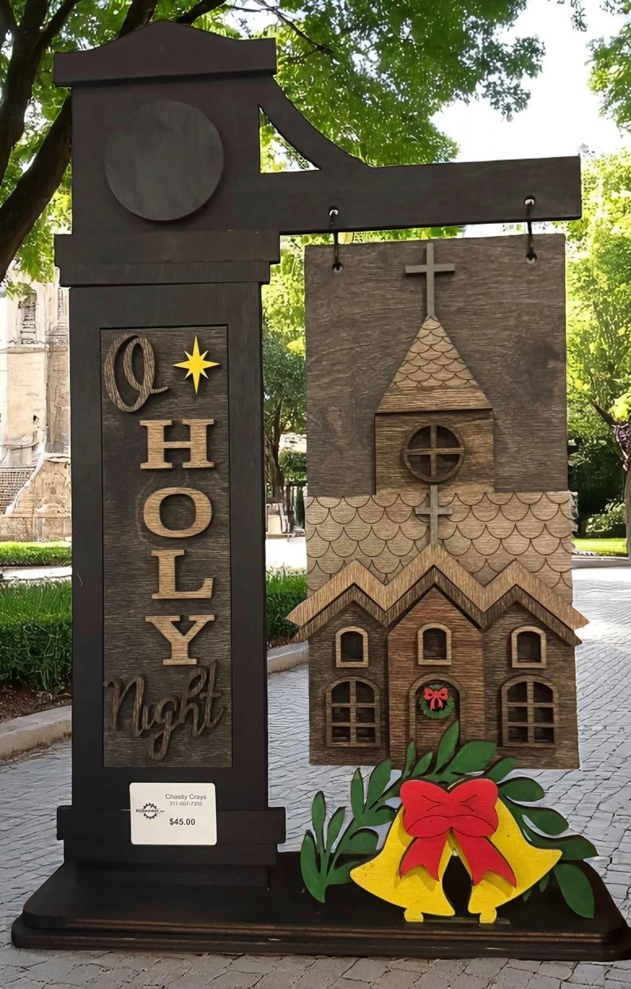 Wooden Christmas decoration featuring a church with a steeple and cross, and a sign that reads "Oh Holy Night" with a yellow star, placed outdoors among trees.