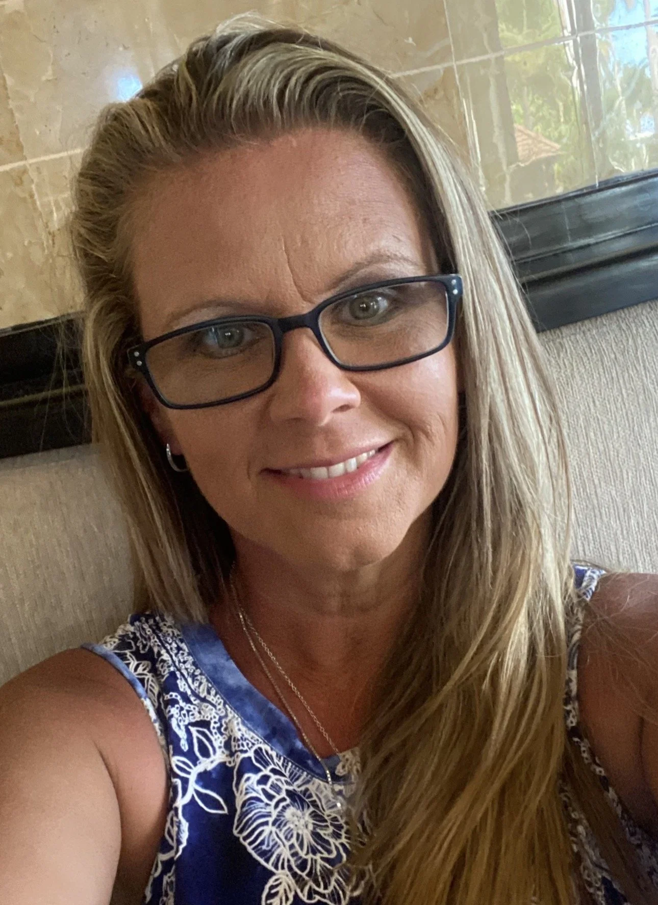 A woman with long blonde hair, glasses, and a blue and white patterned sleeveless top, smiling at the camera indoors, with a beige wall and window in the background.
