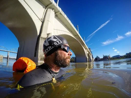Man in swimming gear and sunglasses wading in water near a bridge on a sunny day.