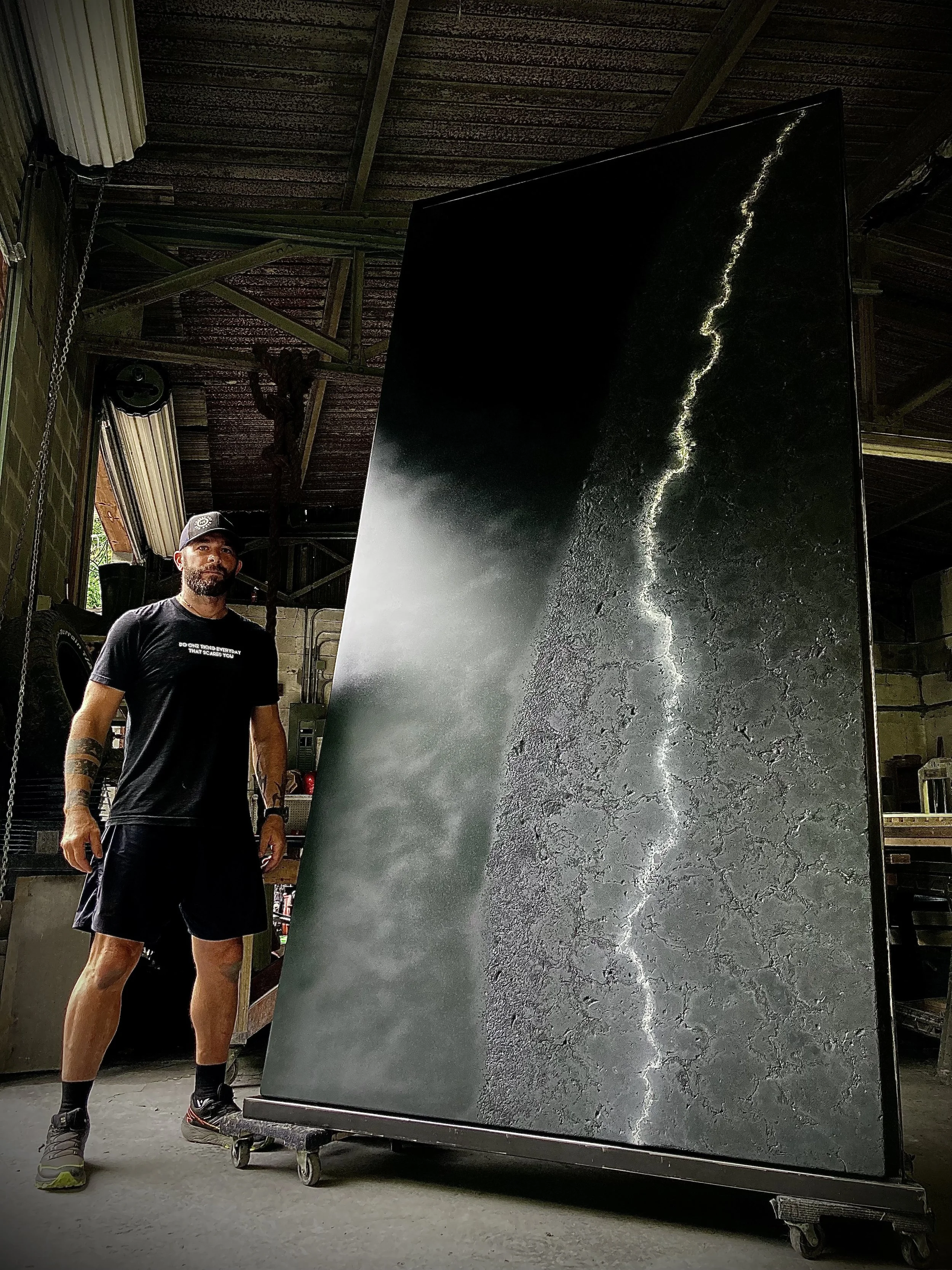 A man with a beard, wearing black shorts, a black t-shirt, sneakers, and a cap, standing next to a large vertical screen displaying a dramatic image of a lightning strike against a dark, stormy sky and cracked earth.