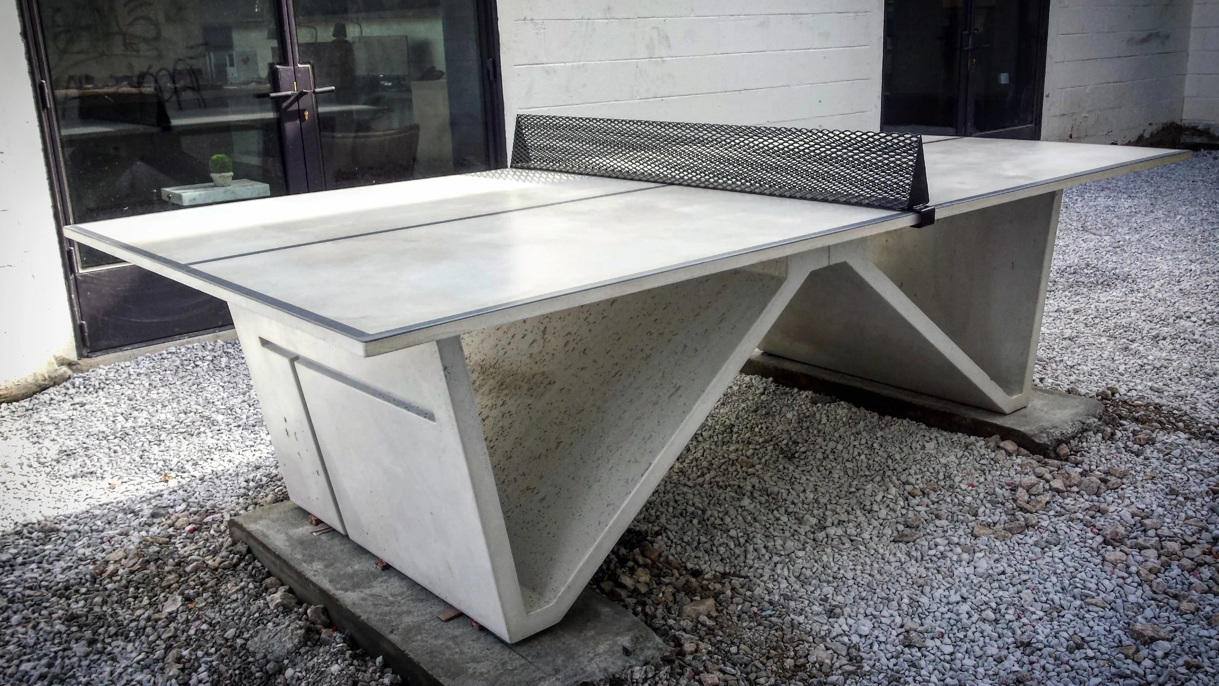 Outdoor ping pong table with a metal net, set on a concrete base and gravel ground near a building with large glass doors.