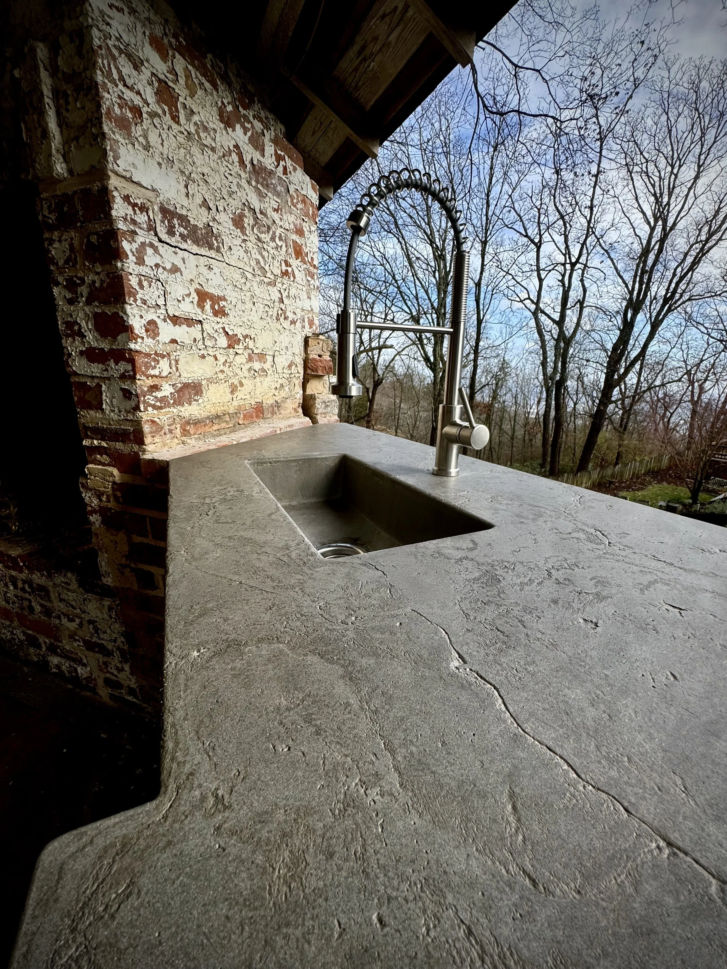 Outdoor kitchen sink with a gray concrete countertop, a modern metal faucet, and a brick wall background on a deck with trees and a blue sky in the distance.