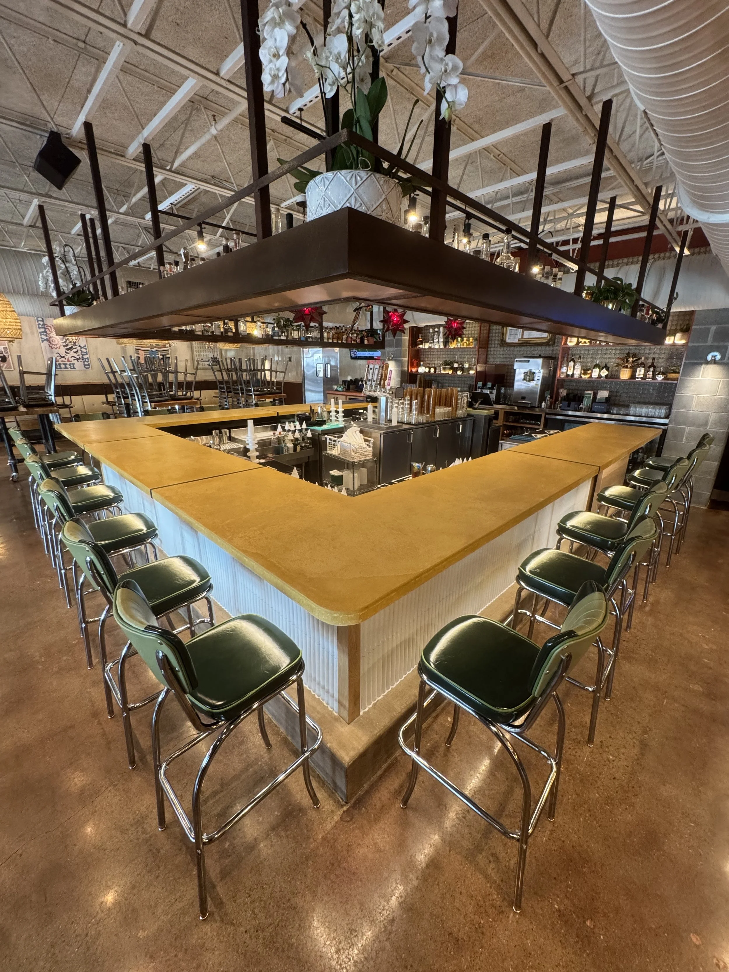 Empty U-shaped bar counter in a restaurant with bar stools, overhead shelf with plants and orchids, and industrial-style decor.