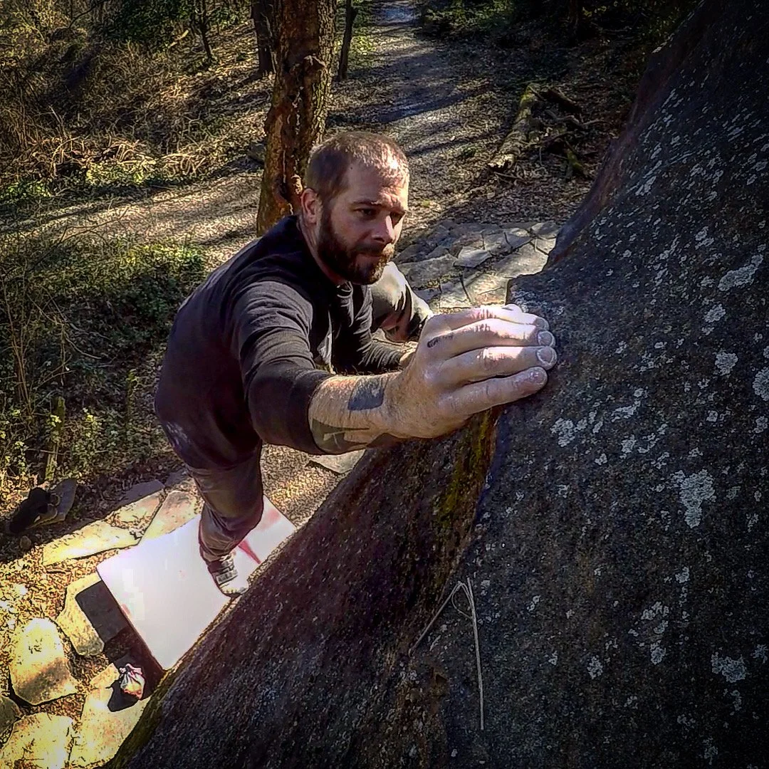 A man rock climbing outdoors on a large boulder, gripping the rock with his hands, with trees and a dirt trail in the background.