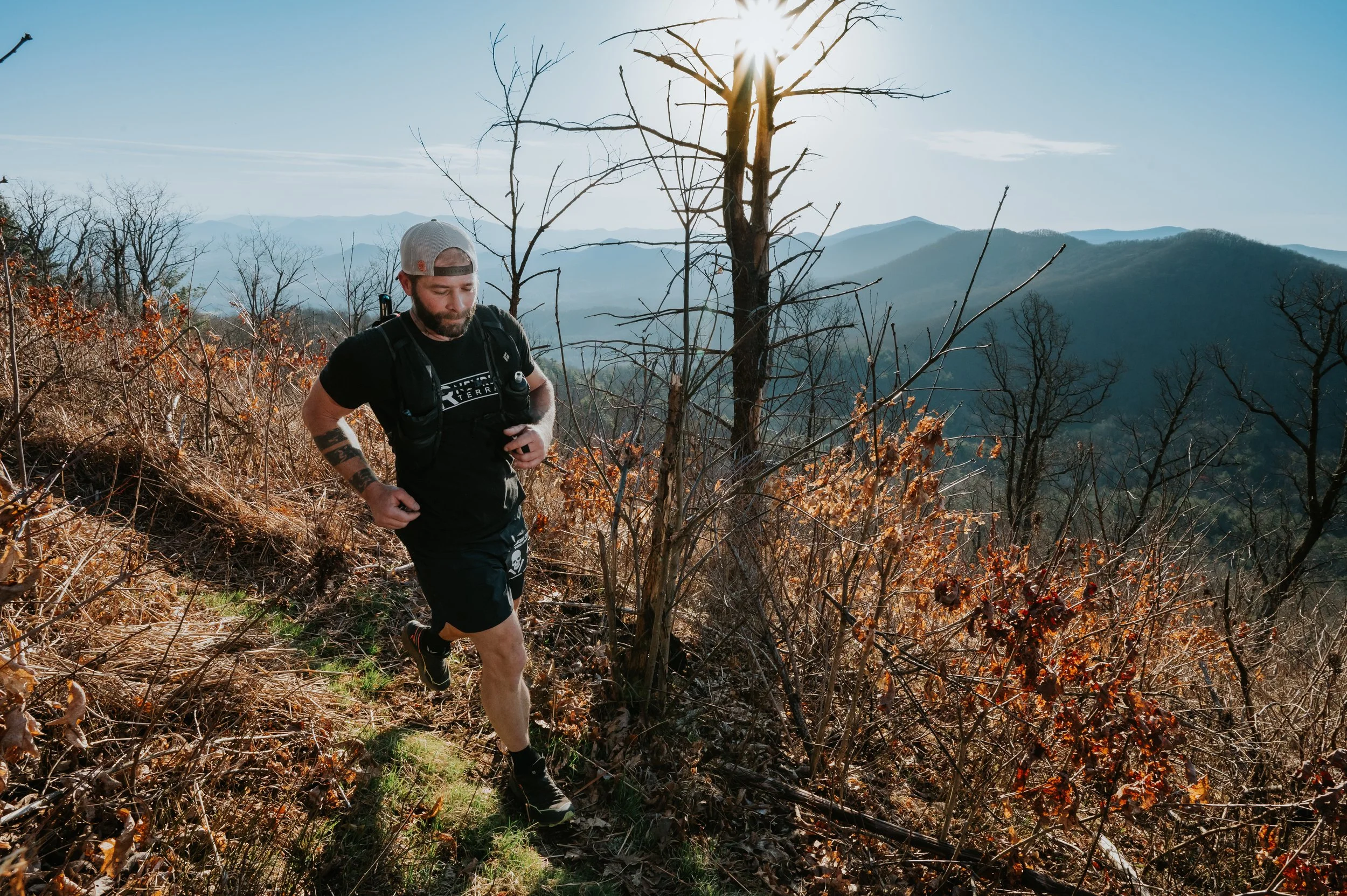 A man trail running on a narrow dirt path in a mountainous area with dry, leafless trees and a mountain range in the background during daylight.