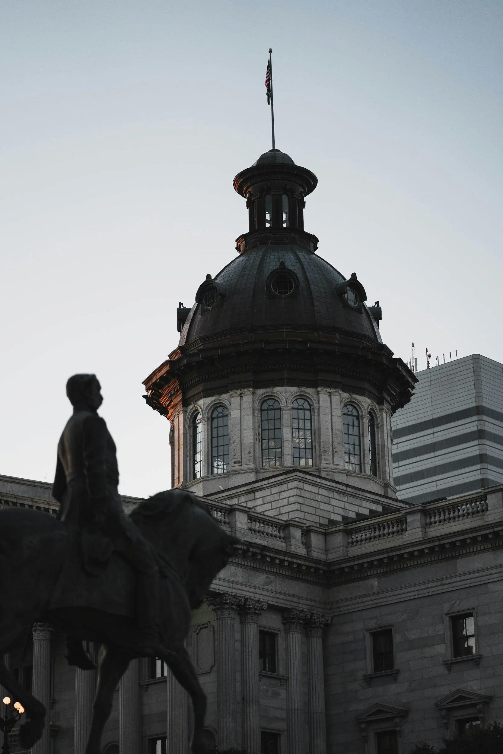 The image features the dome of a historic government building, with a statue of a man on horseback in the foreground. An American flag flies atop the dome. The building has classical architecture with columns and large arched windows. The photo is taken during dusk or dawn.