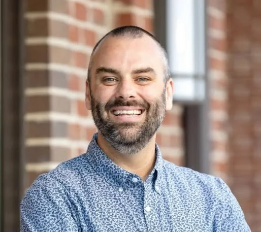 A smiling man with a beard, wearing a blue patterned button-up shirt, standing outside in front of a brick building