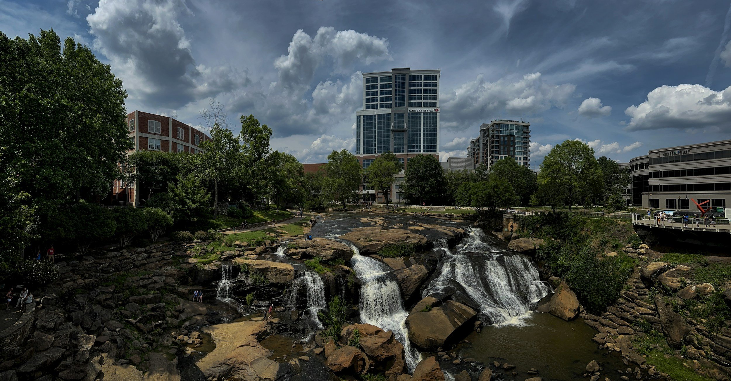 Cityscape with a waterfall flowing over rocks in a park, surrounded by trees, with modern buildings and a cloudy sky in the background.