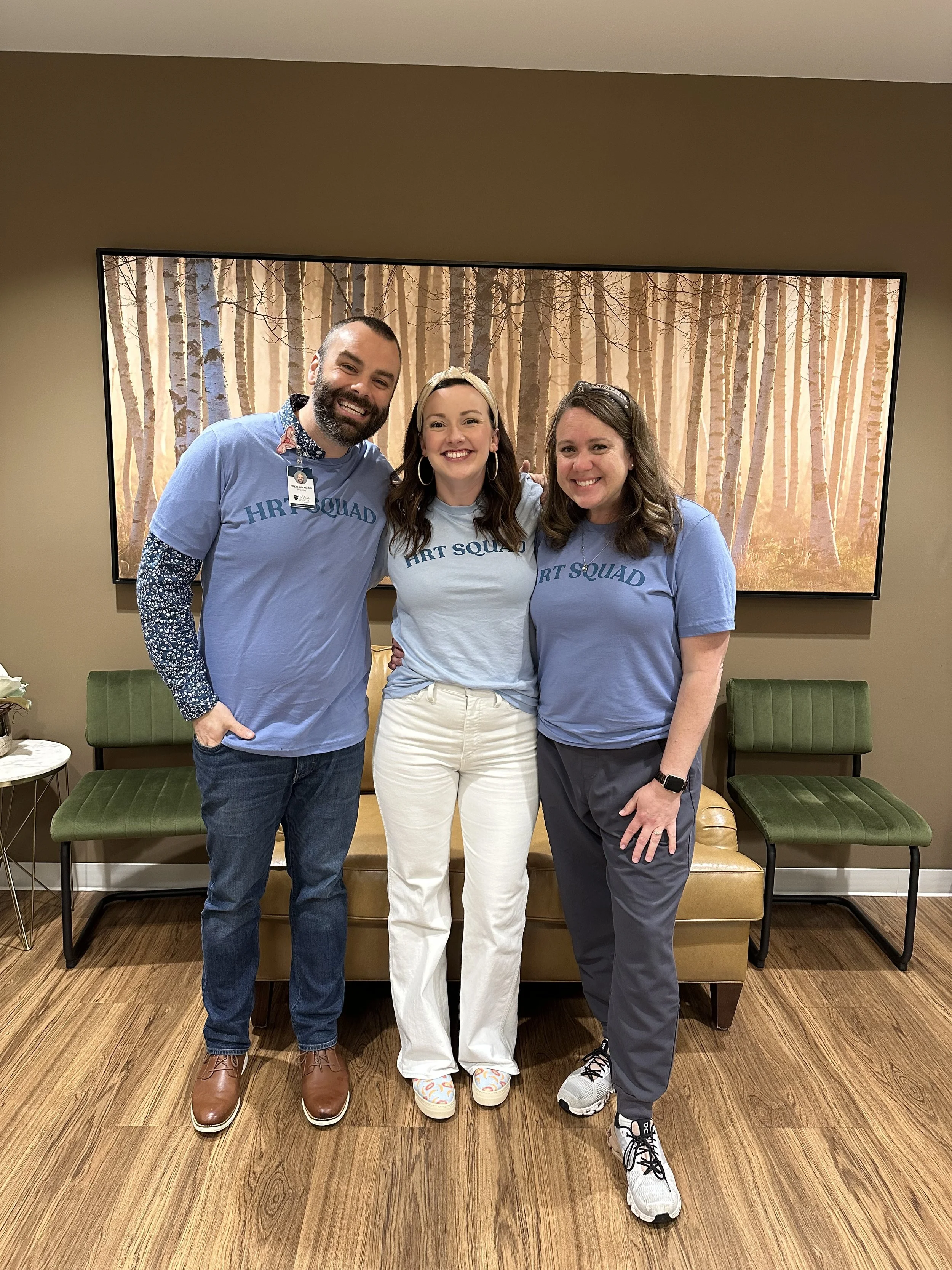 Three people standing together in a room, smiling and wearing matching blue shirts that say 'Art Squad.' There is a forest scene in the background on a screen behind them and two green chairs and a small table nearby.