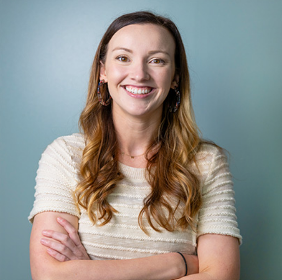 Smiling woman with long curly reddish-brown hair, wearing a cream-colored top and hoop earrings, standing against a blue background.