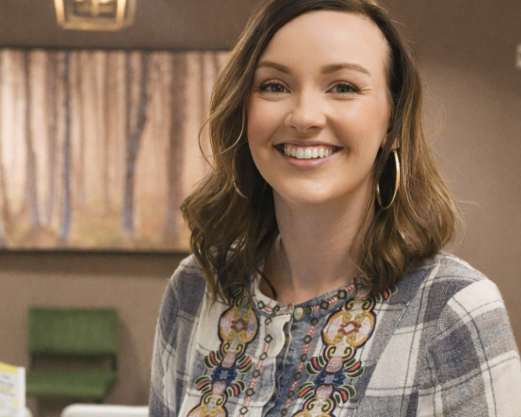 A smiling woman with shoulder-length wavy brown hair, wearing large hoop earrings and a plaid shirt with colorful embroidery, standing indoors with a painting of a forest and curtains in the background.