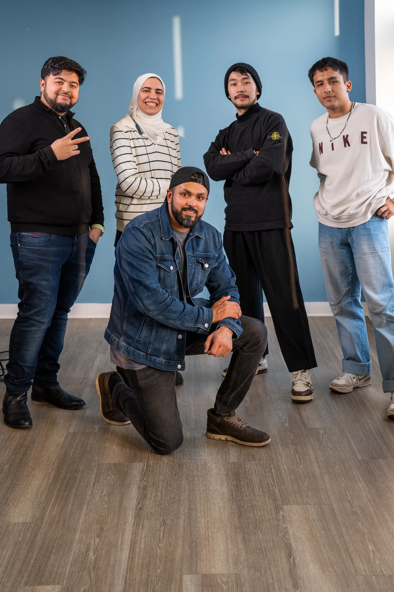 Group of five diverse young adults posing together inside a room with a blue wall and wood flooring, some standing, one kneeling.