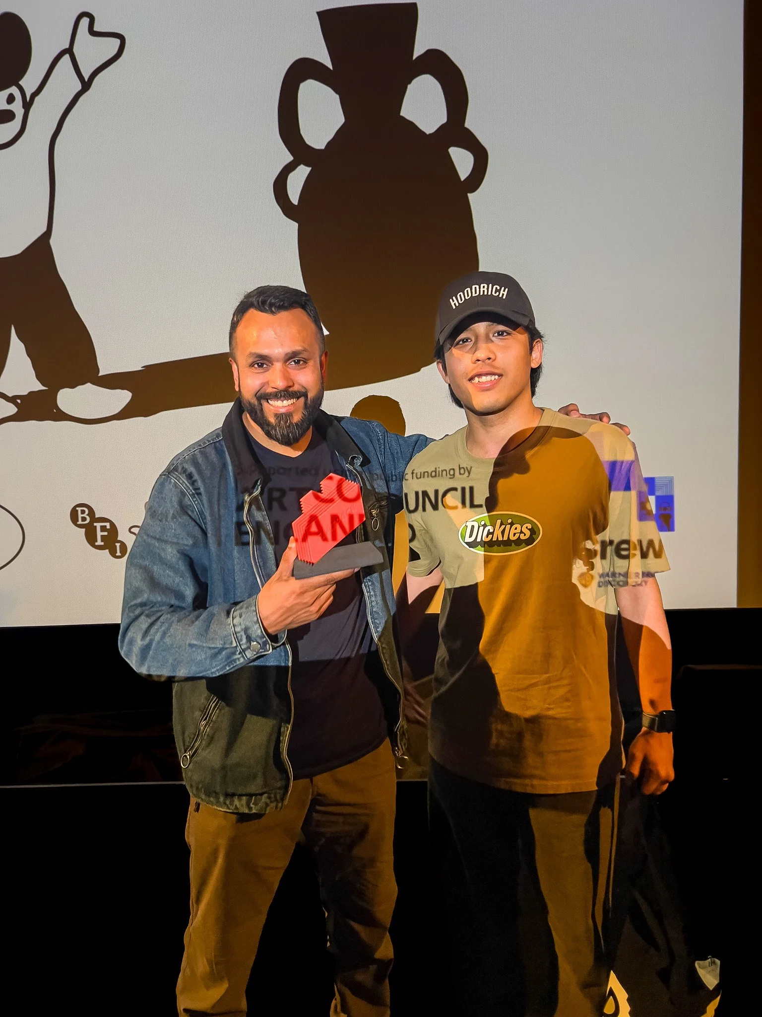 Two men smiling and posing in front of a screen with a drawing of a vase and a character. One man is holding a red sign that says 'U' and 'AND'. The man on the left has a beard and is wearing a denim jacket, while the man on the right is wearing a black Cap with 'HOODRICH' written on it and a t-shirt with a Dickies logo.
