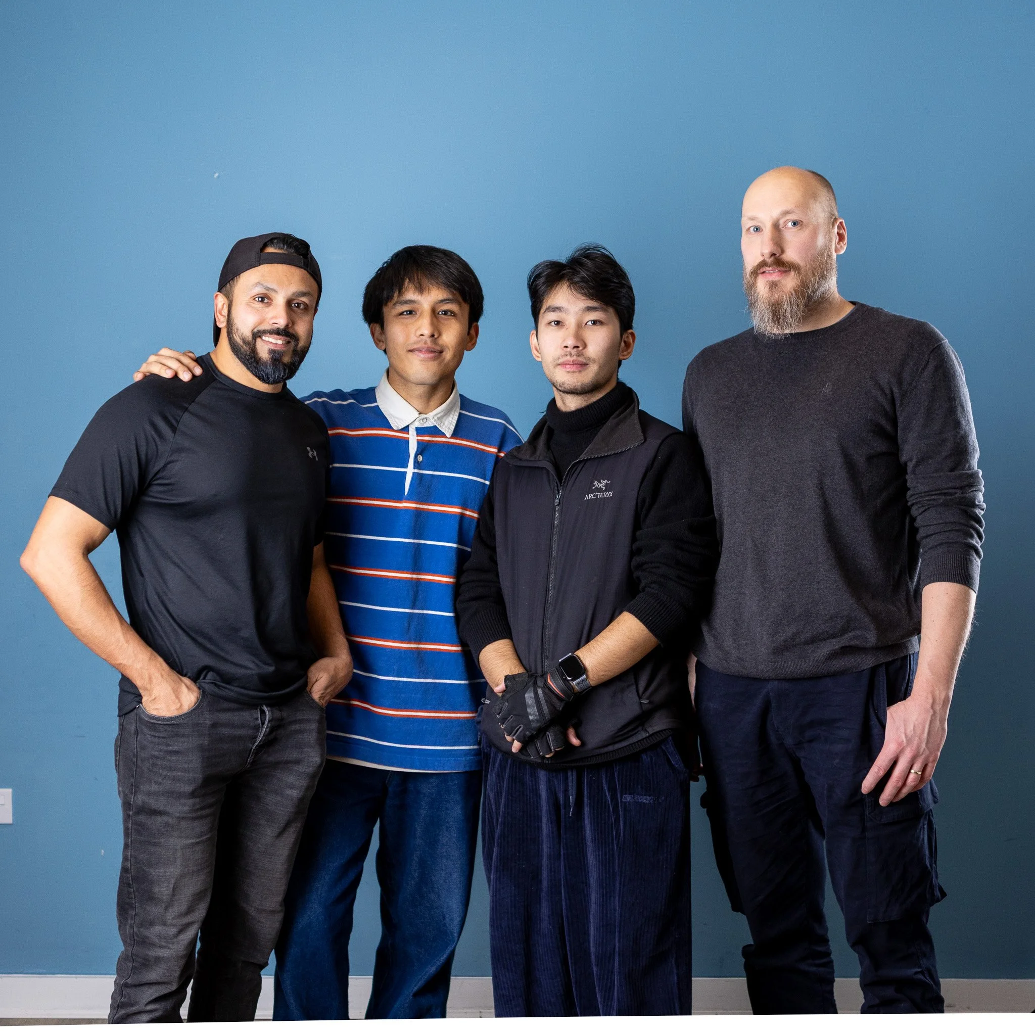Four men standing together against a blue background, smiling and looking at the camera.