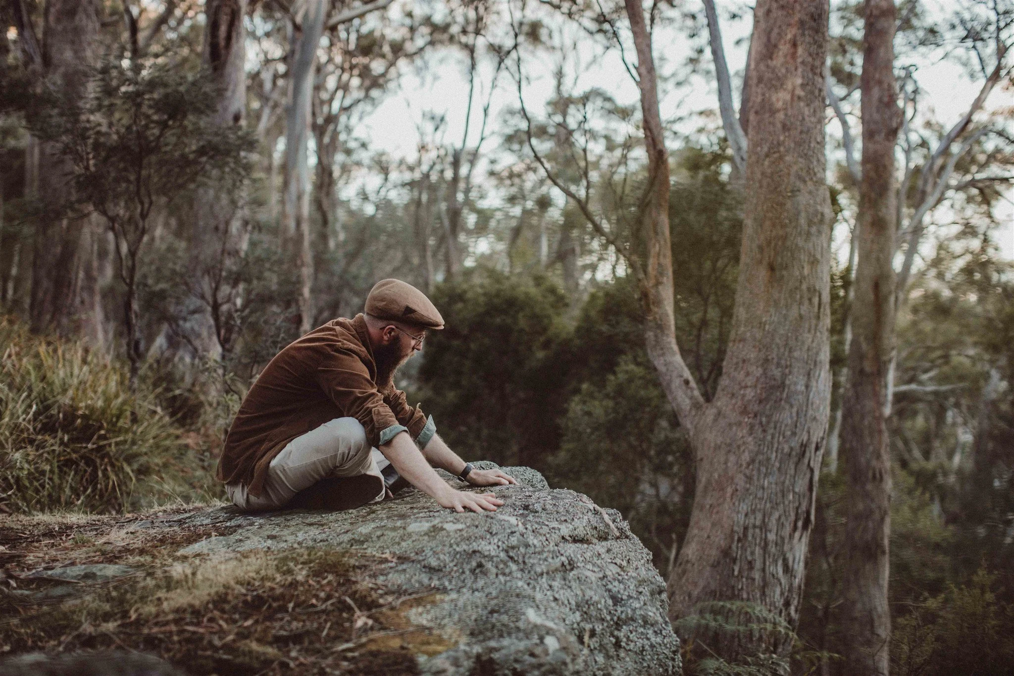 A man with glasses, a brown hat, and brown jacket crouches on a rock in a forested area, reaching out with his hand, surrounded by tall trees and greenery.