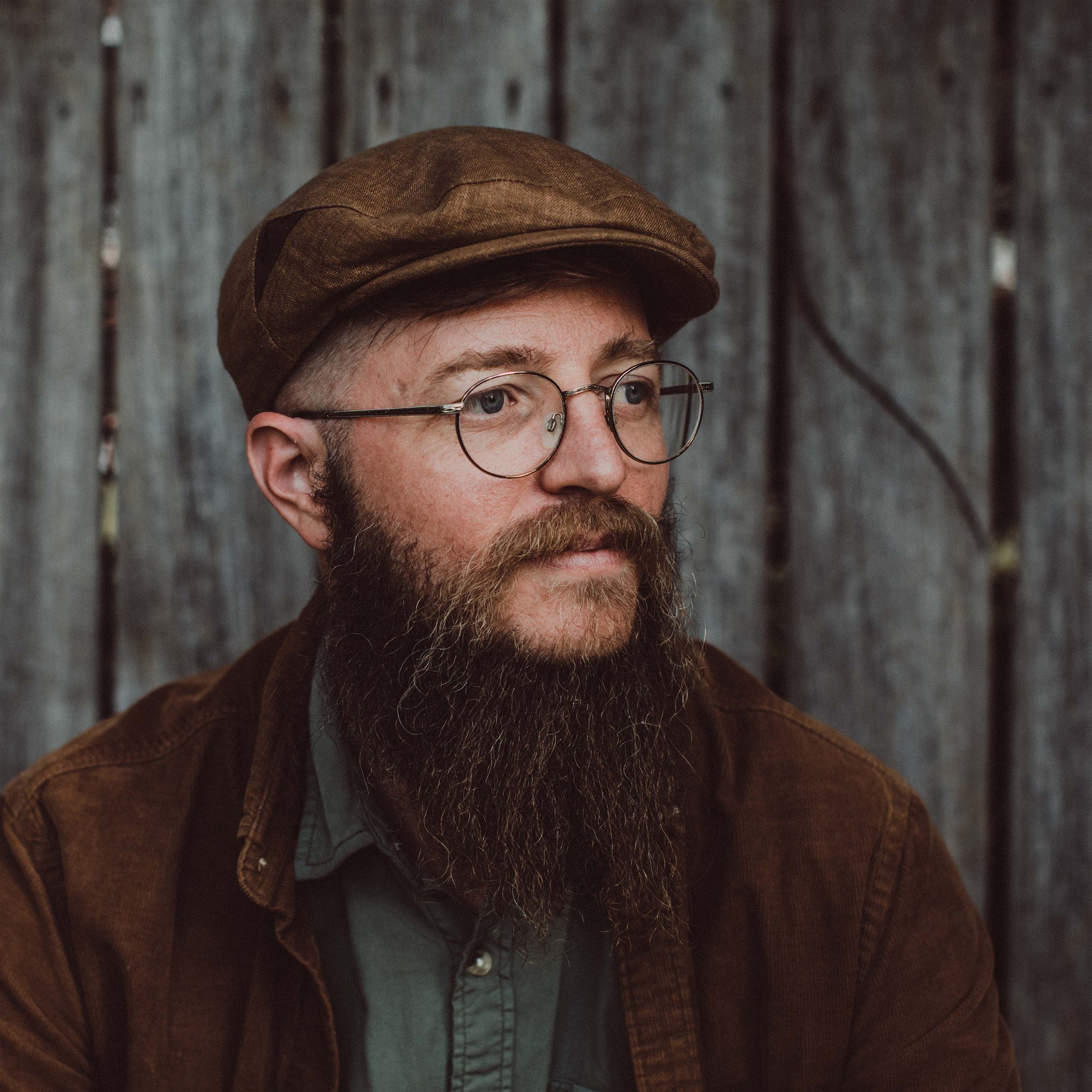 Close-up of a man's face showing part of his forehead, eye, glasses, beard, and neck, wearing a textured brown cap and a brown jacket with a gray top underneath, against a background of weathered wooden planks.