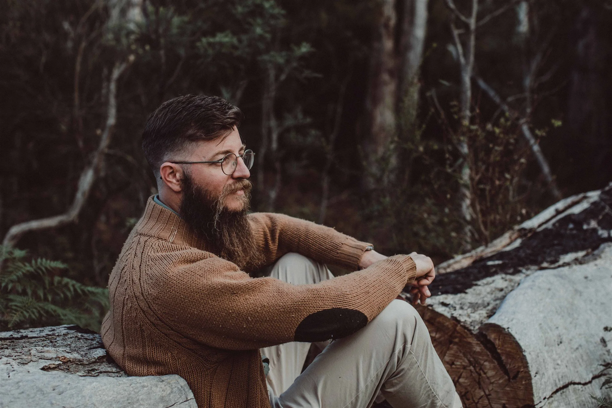 A man with glasses, a beard, and brown hair, wearing a brown sweater and beige pants, sitting outdoors on a log near a forest, looking to the side.