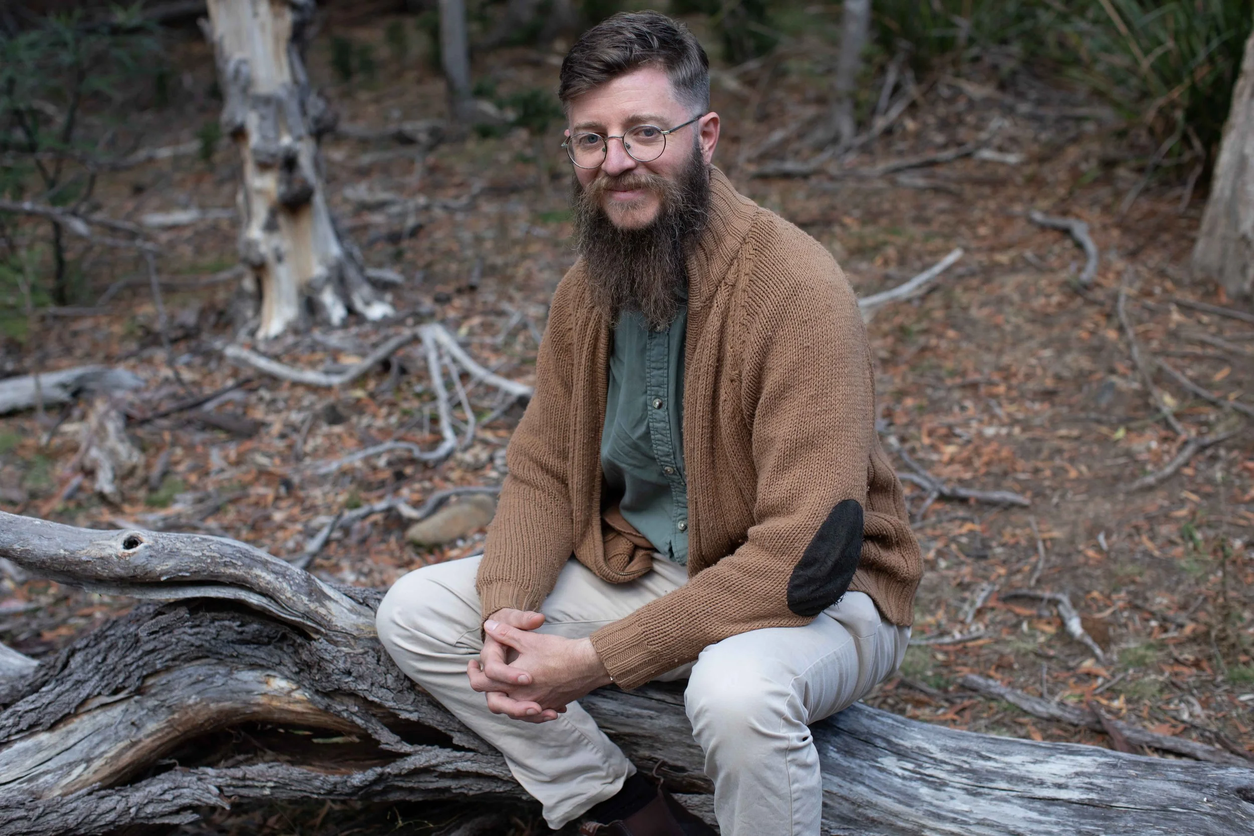 A man with glasses and a beard, wearing a brown sweater and light-colored pants, sitting on a fallen tree in a forest during daytime.