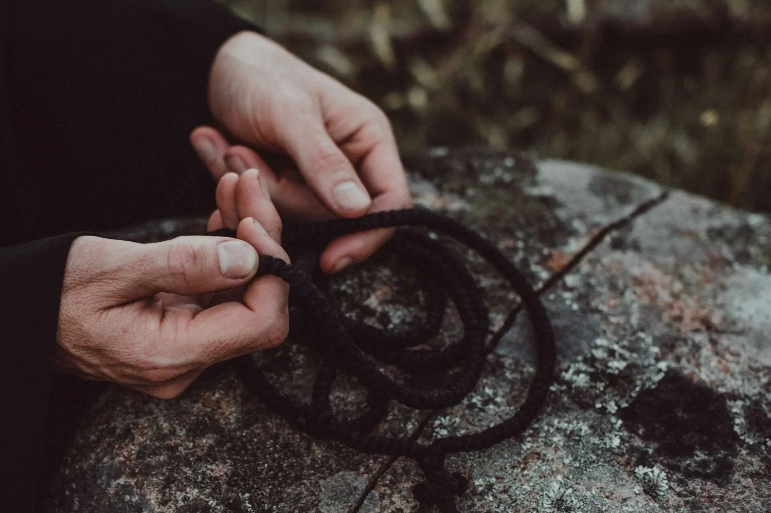Person tying black prayer rope on a large, rough granite rock outdoors.
