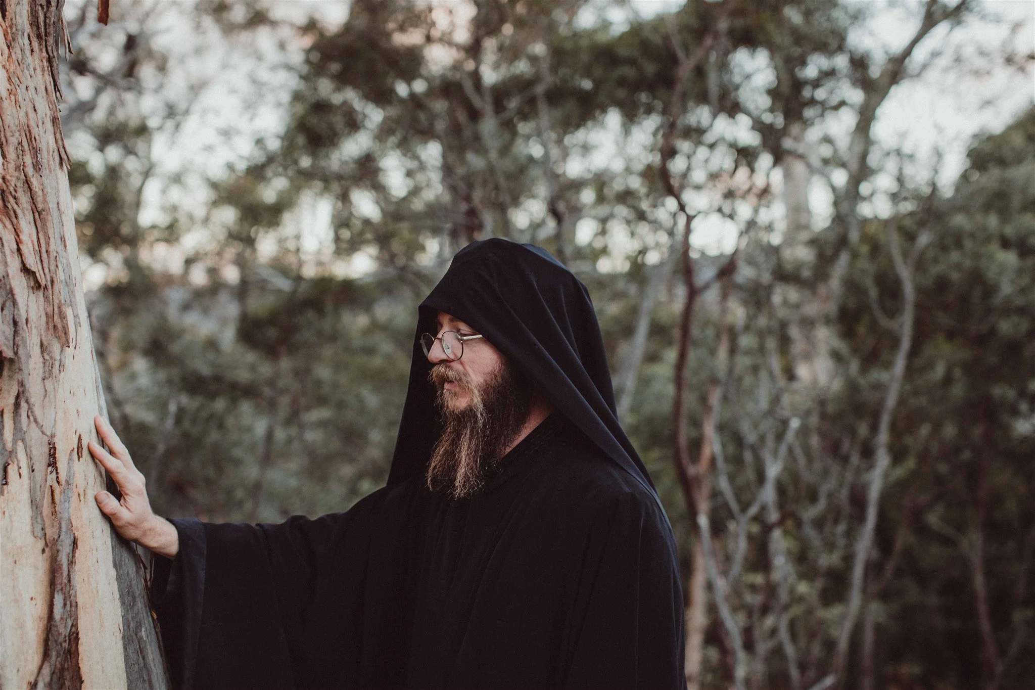 A man with a beard wearing glasses and a black hooded cloak touches a tree in a forest.