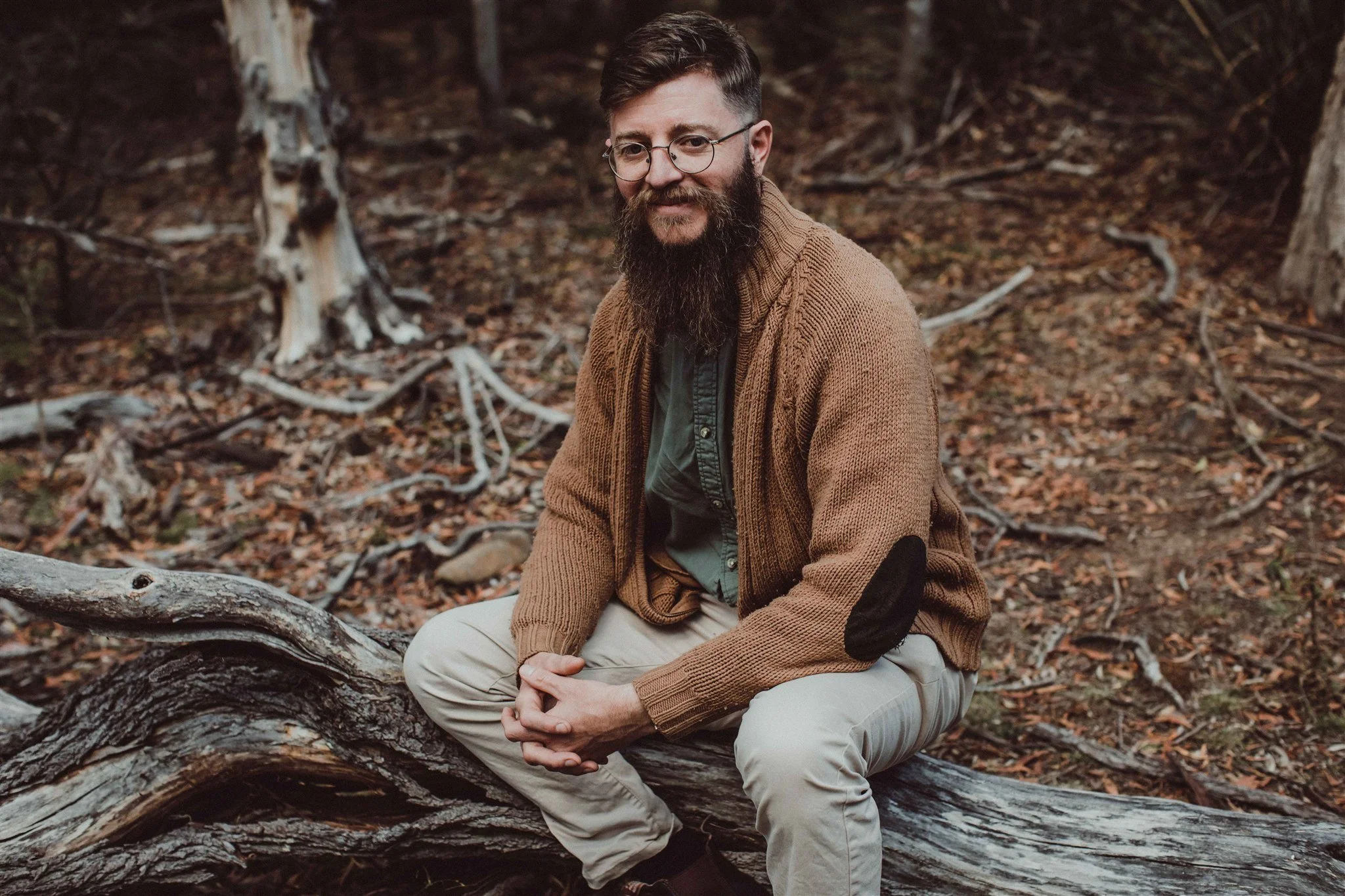 A man with glasses and a beard sitting on a fallen tree in a forest with brown leaves and trees in the background.