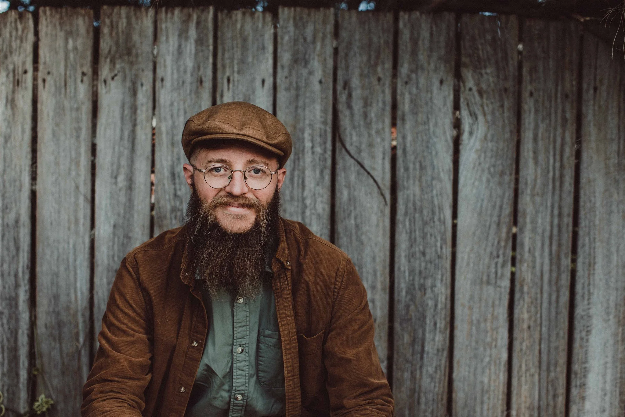 A man with glasses, a brown beard, and a brown flat cap, wearing a brown jacket, sitting outdoors in front of a wooden fence.