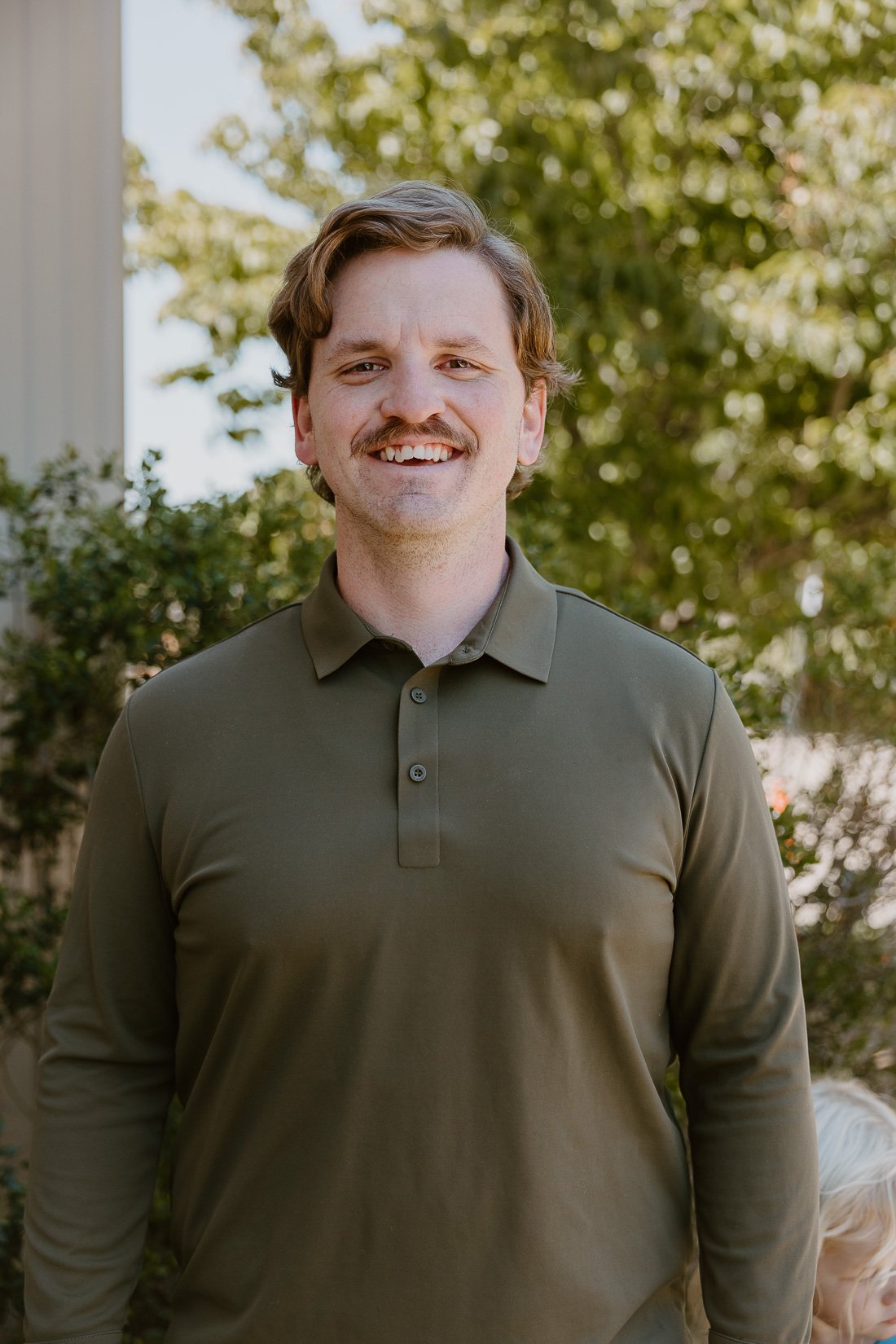 A smiling man with light skin, brown hair, a mustache, wearing an olive green collared shirt, standing outdoors with trees and a building in the background.