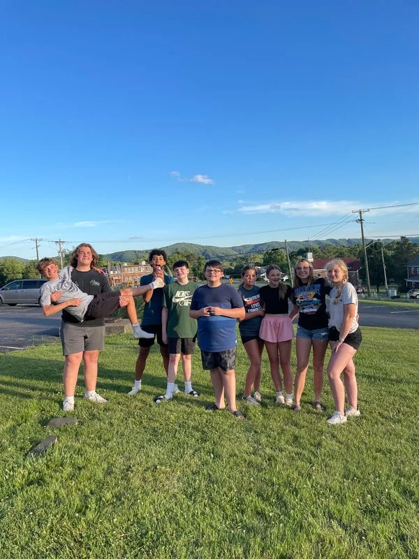 Group of nine teenagers and children standing on grass outdoors under a clear blue sky, with hills and buildings in the background, one girl being held horizontally by a boy.