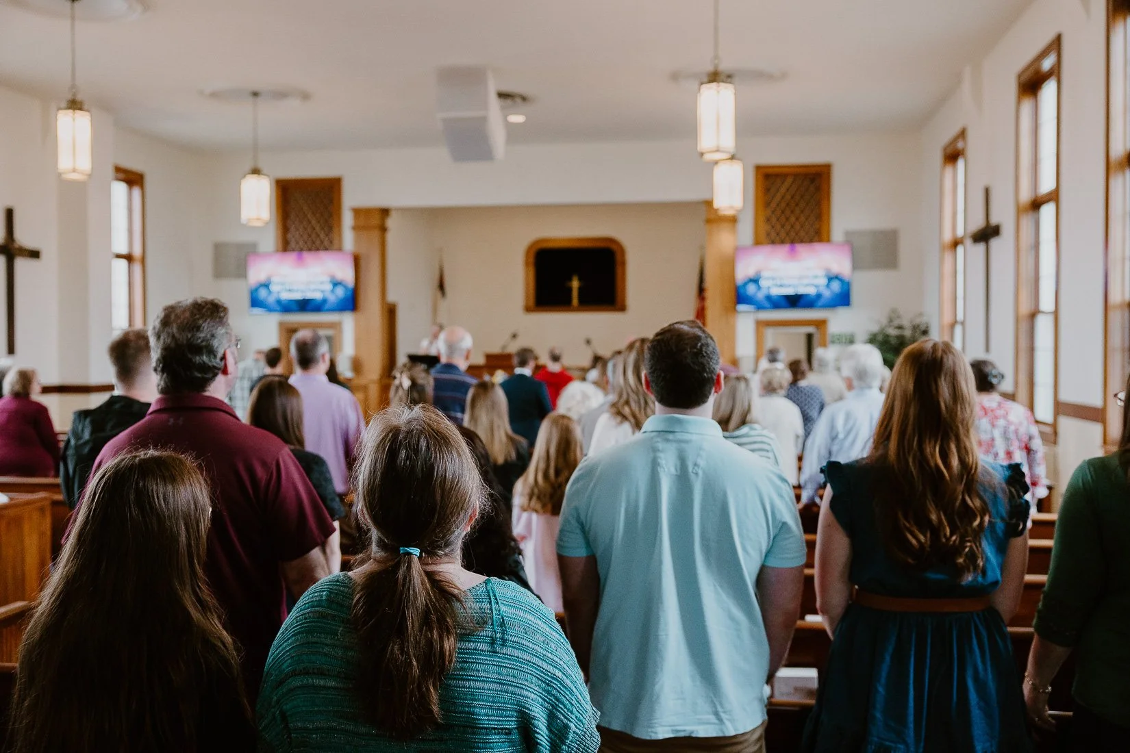 A diverse group of people standing inside a church during a service, facing the altar with a cross, two screens displaying lyrics or hymns, and wooden pews.