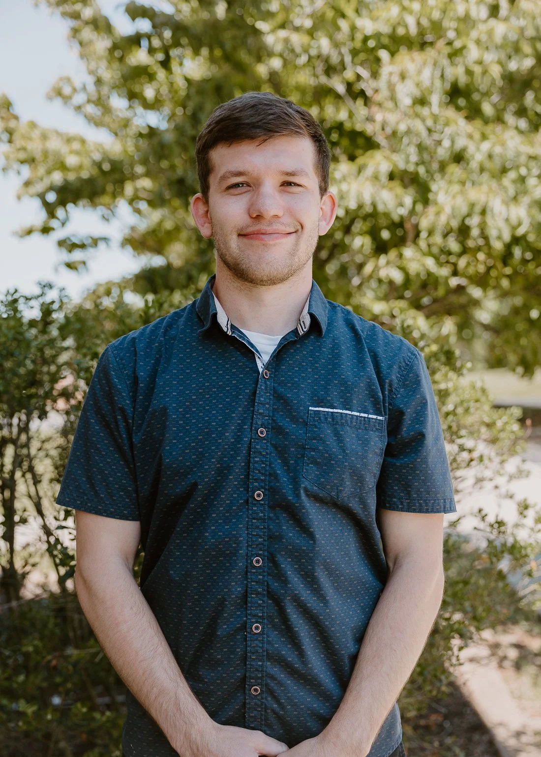 A young man with short brown hair, wearing a dark blue short-sleeved button-up shirt with a subtle pattern, stands outdoors smiling against a background of trees and greenery.