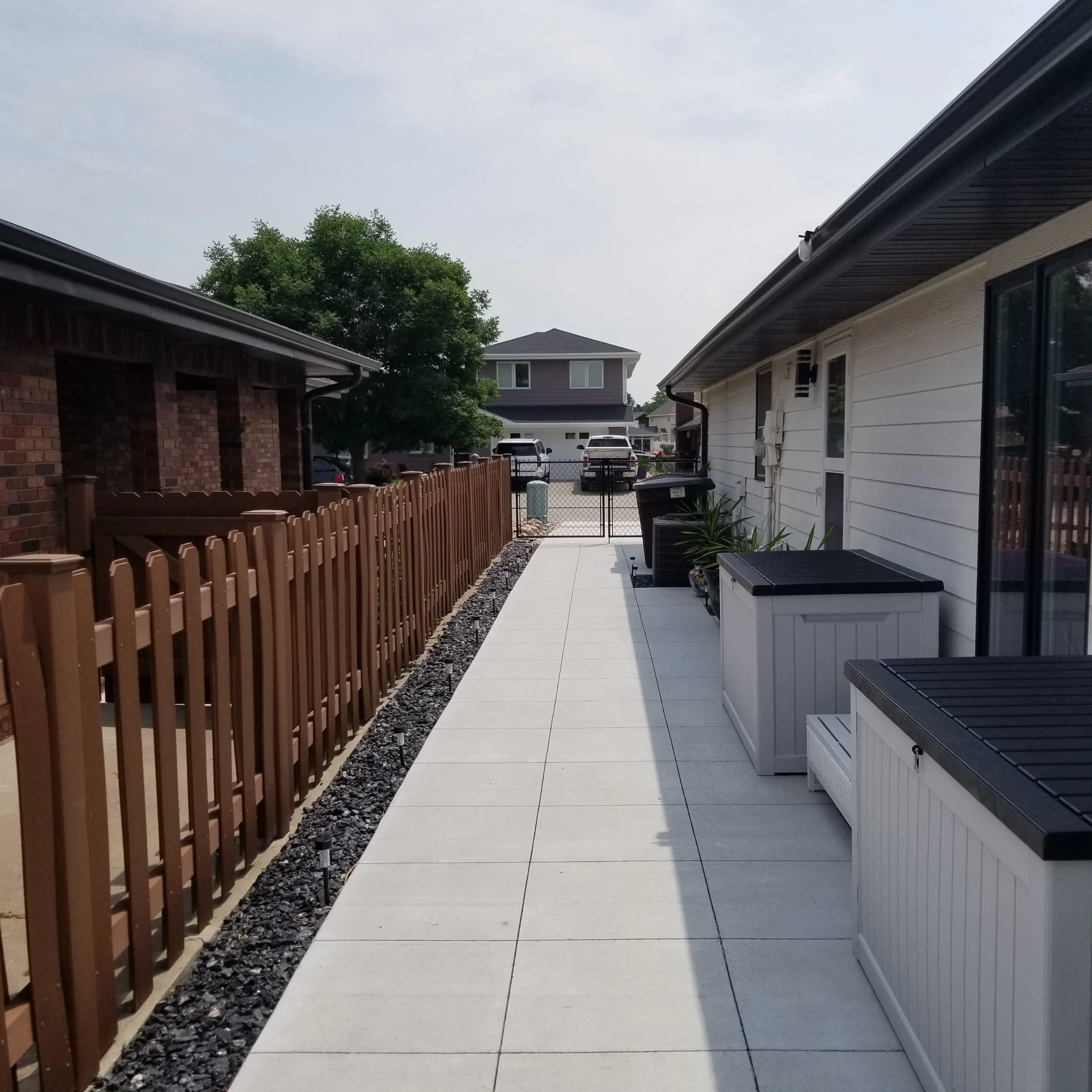 A backyard patio with white concrete tiles, a brown wooden fence on the left, and a white house with gardening storage on the right. There are plants and a manhole cover near the house, and in the background, there is a street with parked cars, trees