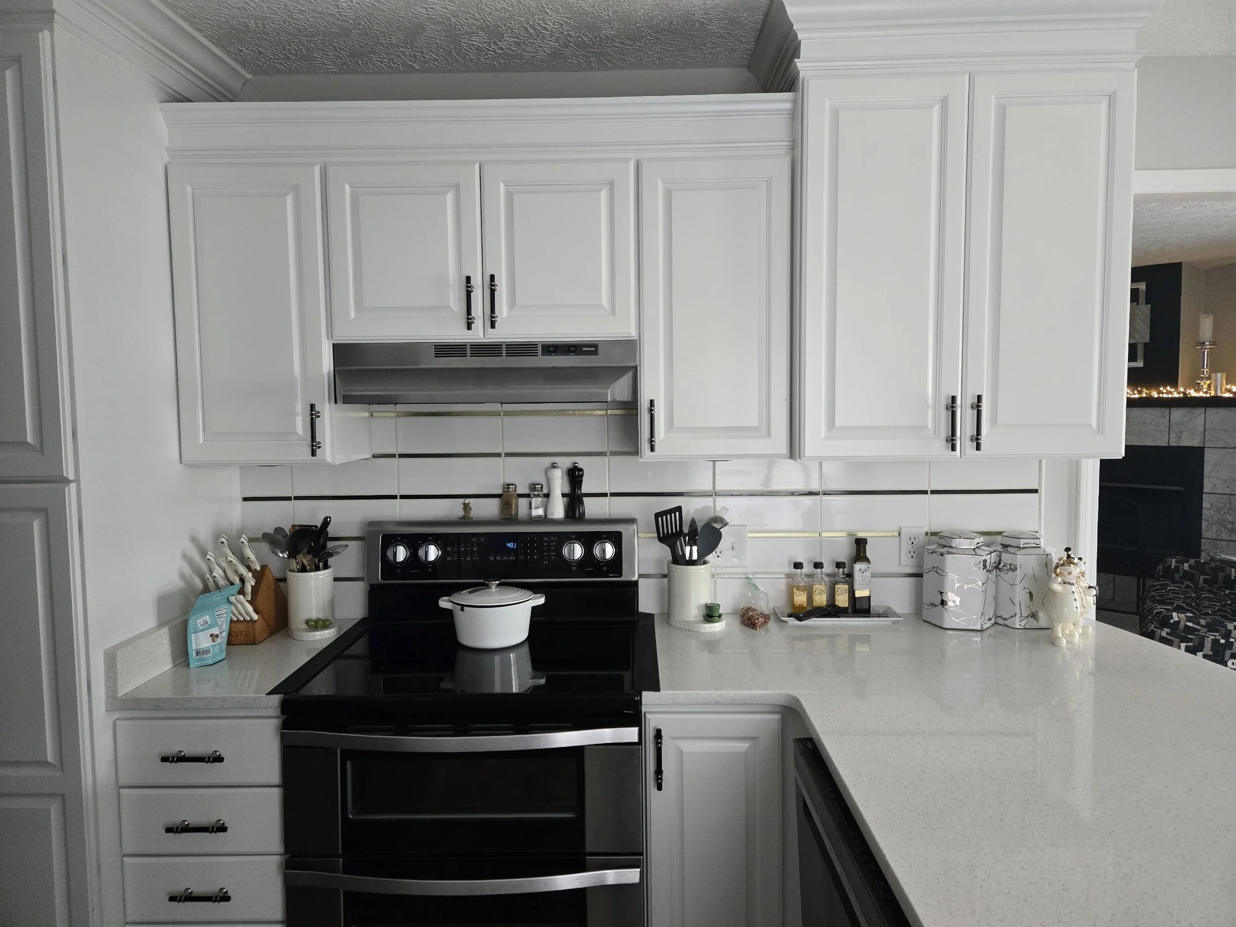 Modern kitchen with white cabinets, black stove with a white pot, and a white countertop with various small kitchen items and decorations.