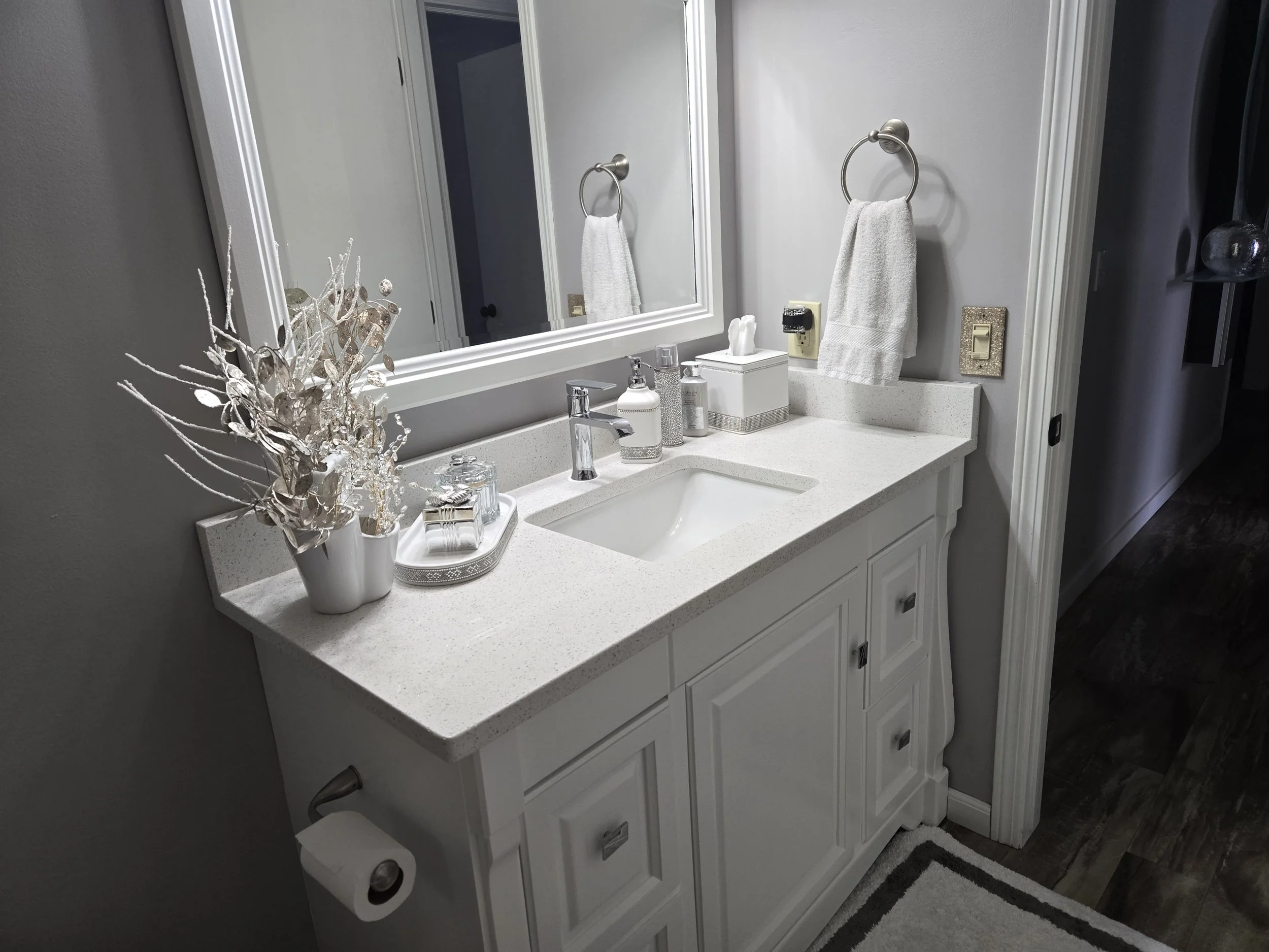 Bathroom vanity with white countertop, mirror, towel ring with white towel, decorative vase, and various toiletries, with a reflection of the opposite wall.