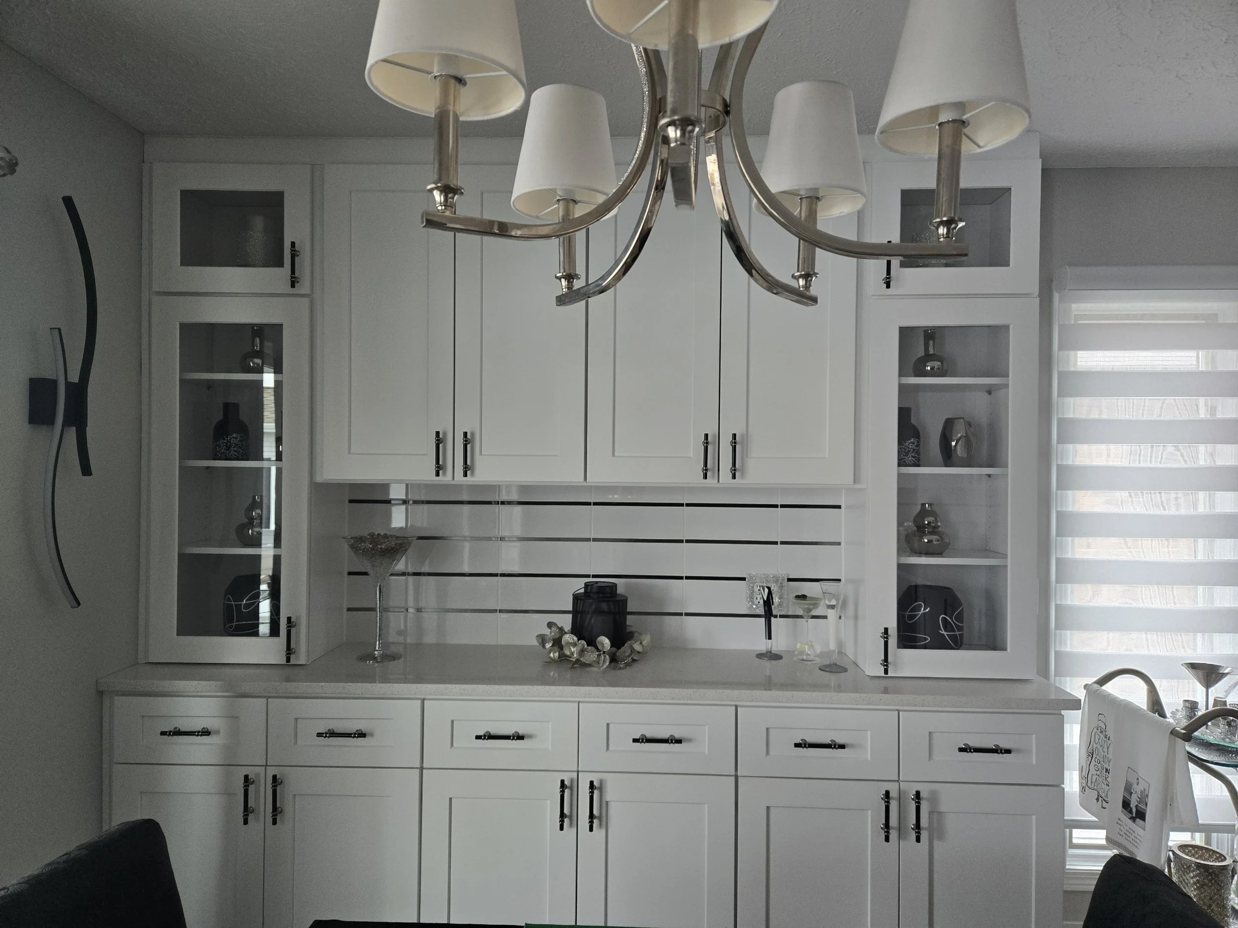 White kitchen cabinet with glass doors, decorative objects inside, silver handles, a chandelier with lampshades hanging above, and a window with striped shades to the right.