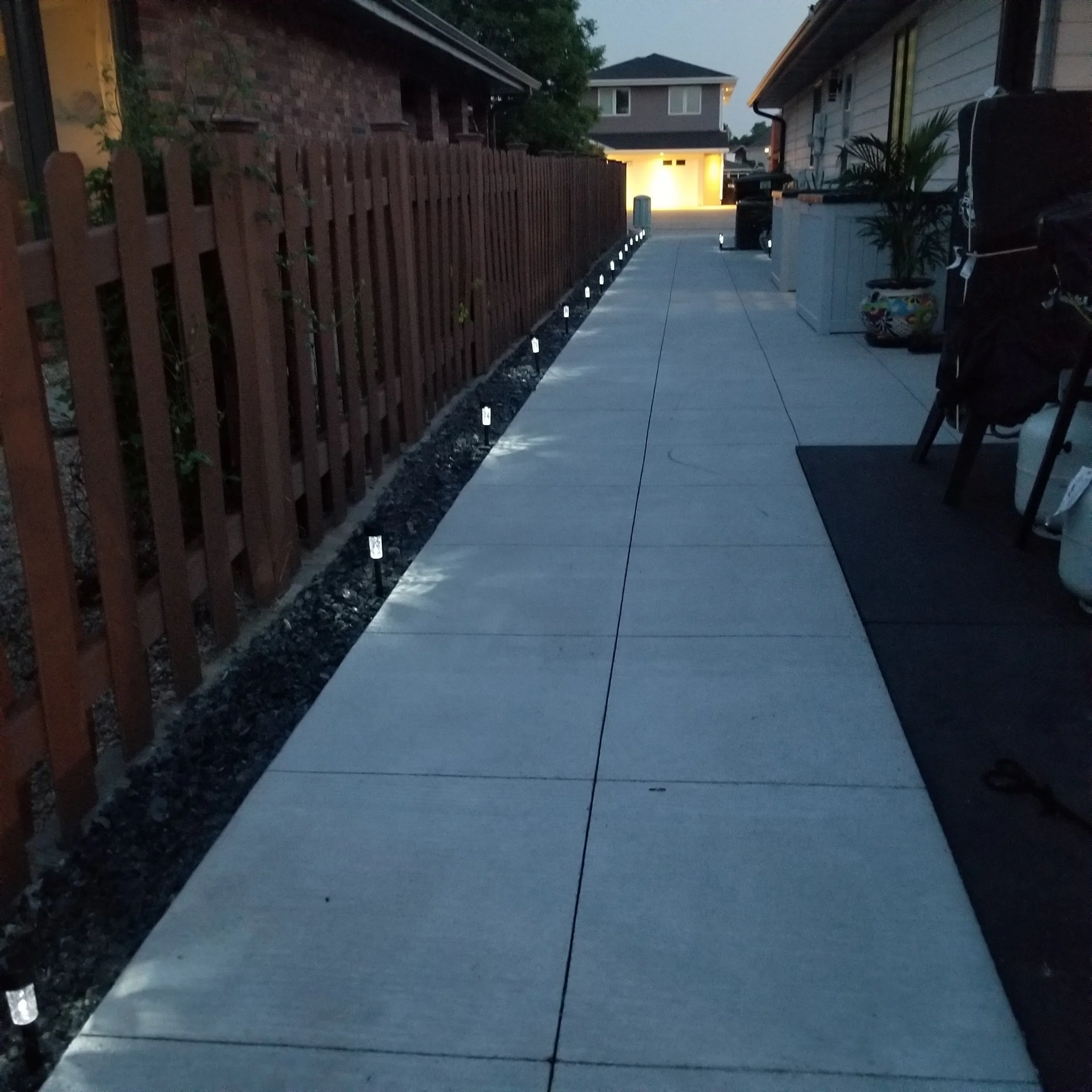 A sidewalk at dusk with small pathway lights along its edge, adjacent to a wooden fence and residential houses.