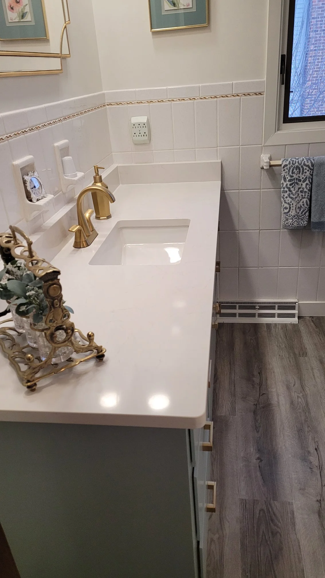 A bathroom vanity with a white countertop and a square sink, gold faucets, a decorative candle holder, a wall-mounted mirror, framed pictures, an outlet, and a window with blinds. Towels are hanging on a rod nearby, and the floor is wood-like.