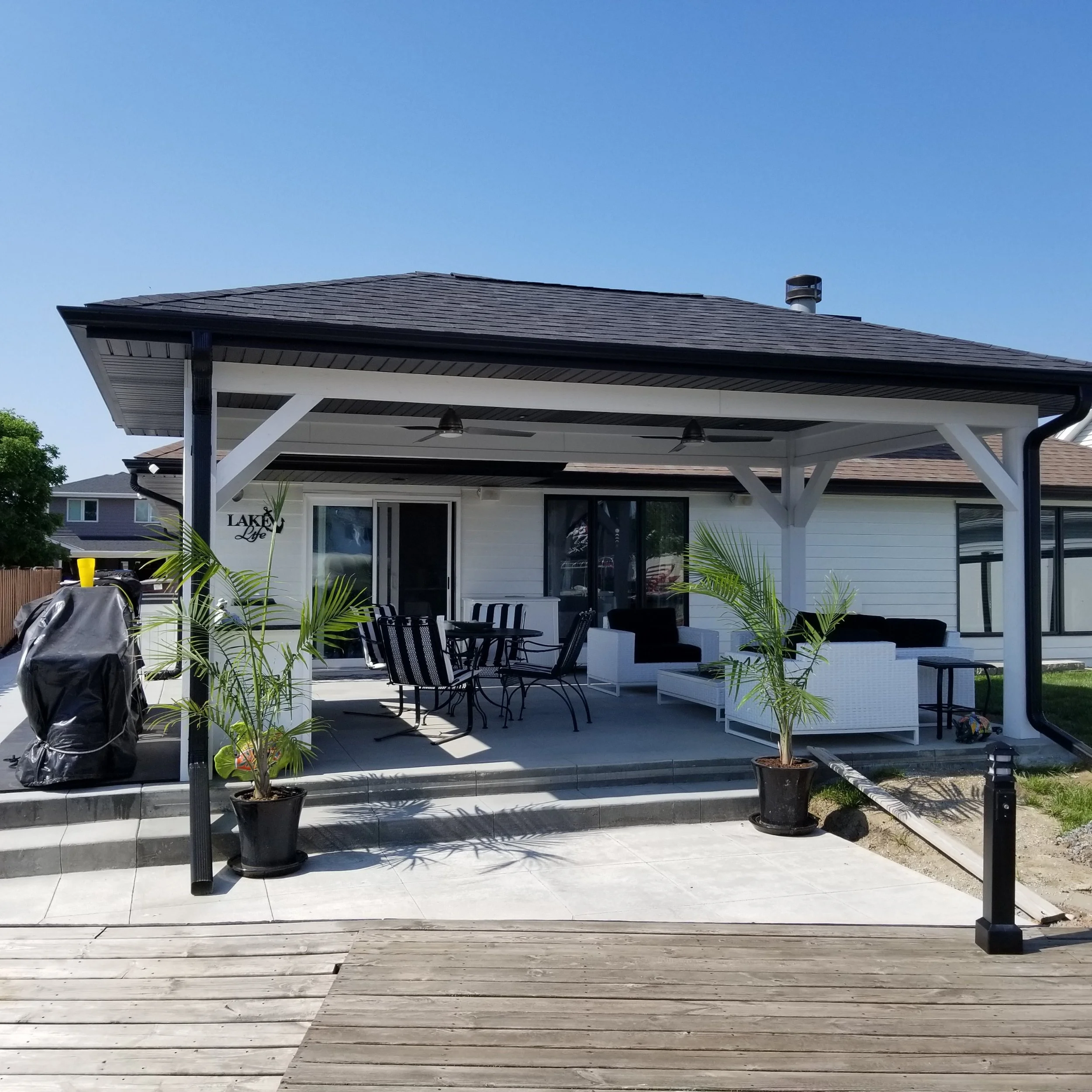 Backyard patio with black metal dining table and chairs, white outdoor sofa with cushions, potted palm trees, and a covered deck area on a sunny day.